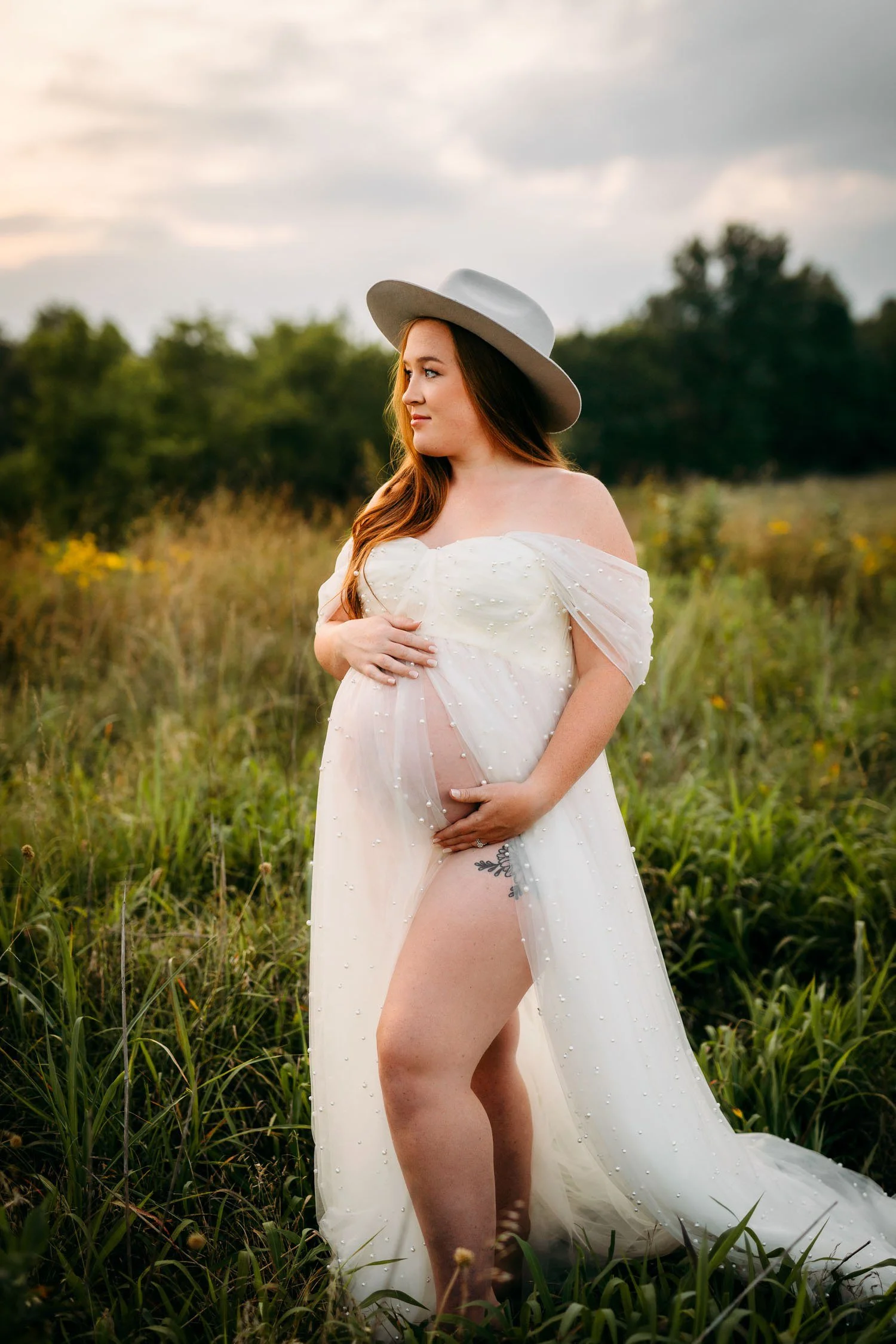 Pregnant woman in a flowing white gown and hat posing in a field during a St. Louis pregnancy photography session.