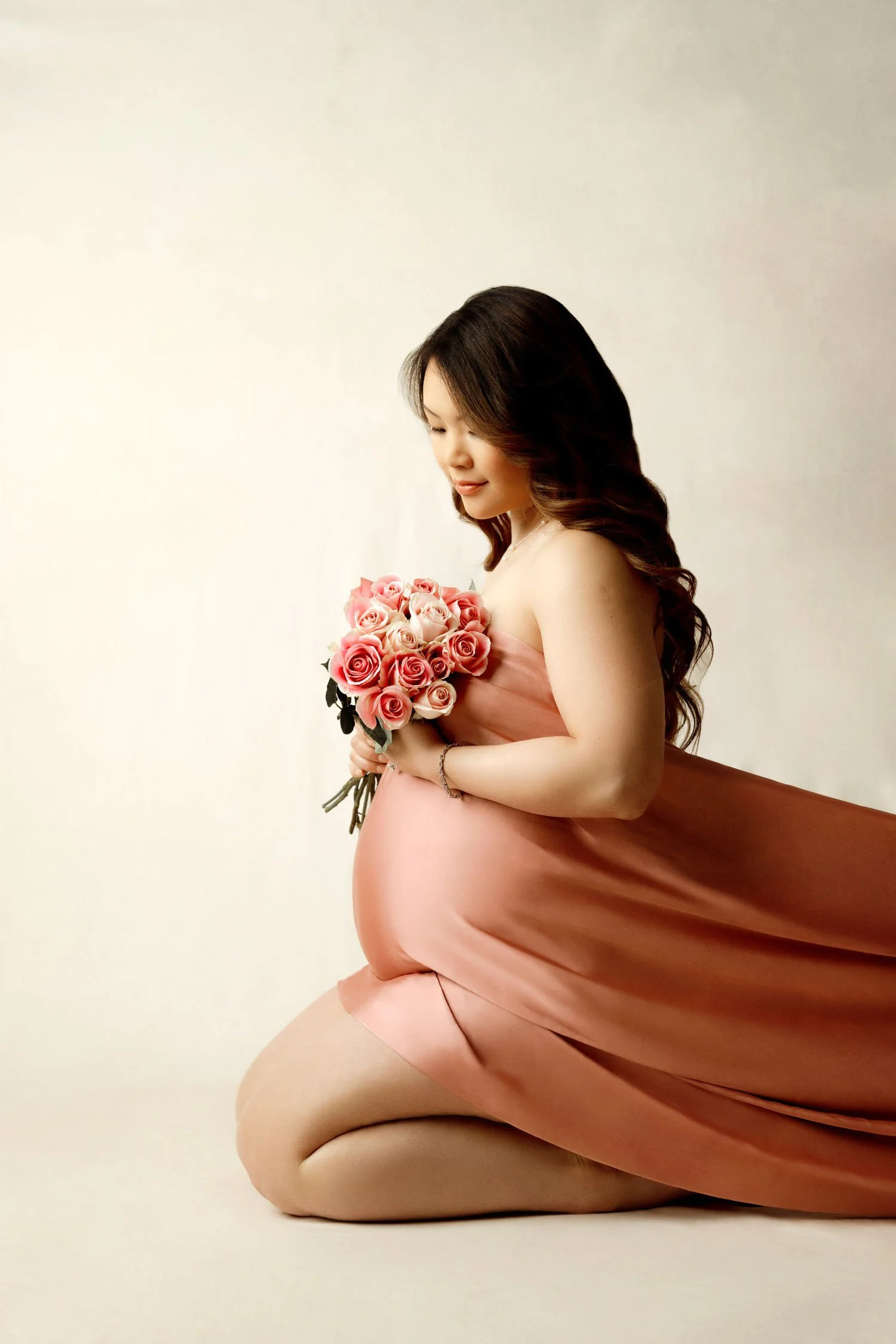 Pregnant woman holding pink flowers while wearing a flowing blush maternity draping satin fabric.