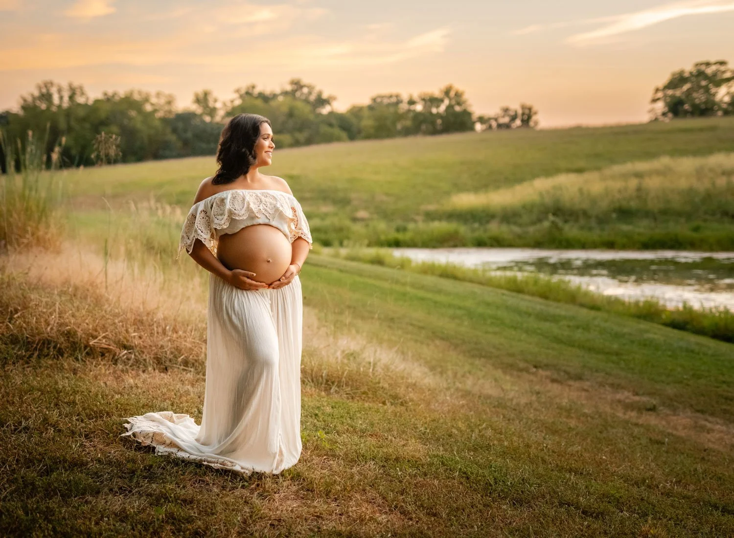 Expecting mother in a white gown standing in a grassy field at sunset during an outdoor maternity portrait.