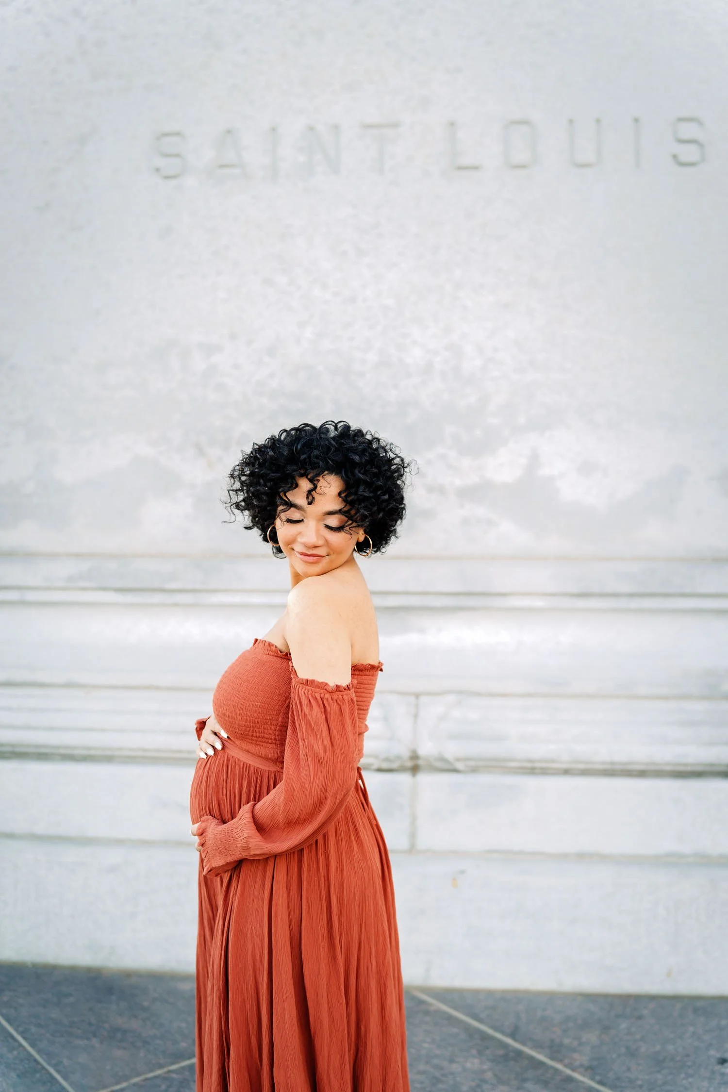 Expecting mother in a rust gown posing outdoors in front of a St. Louis backdrop.