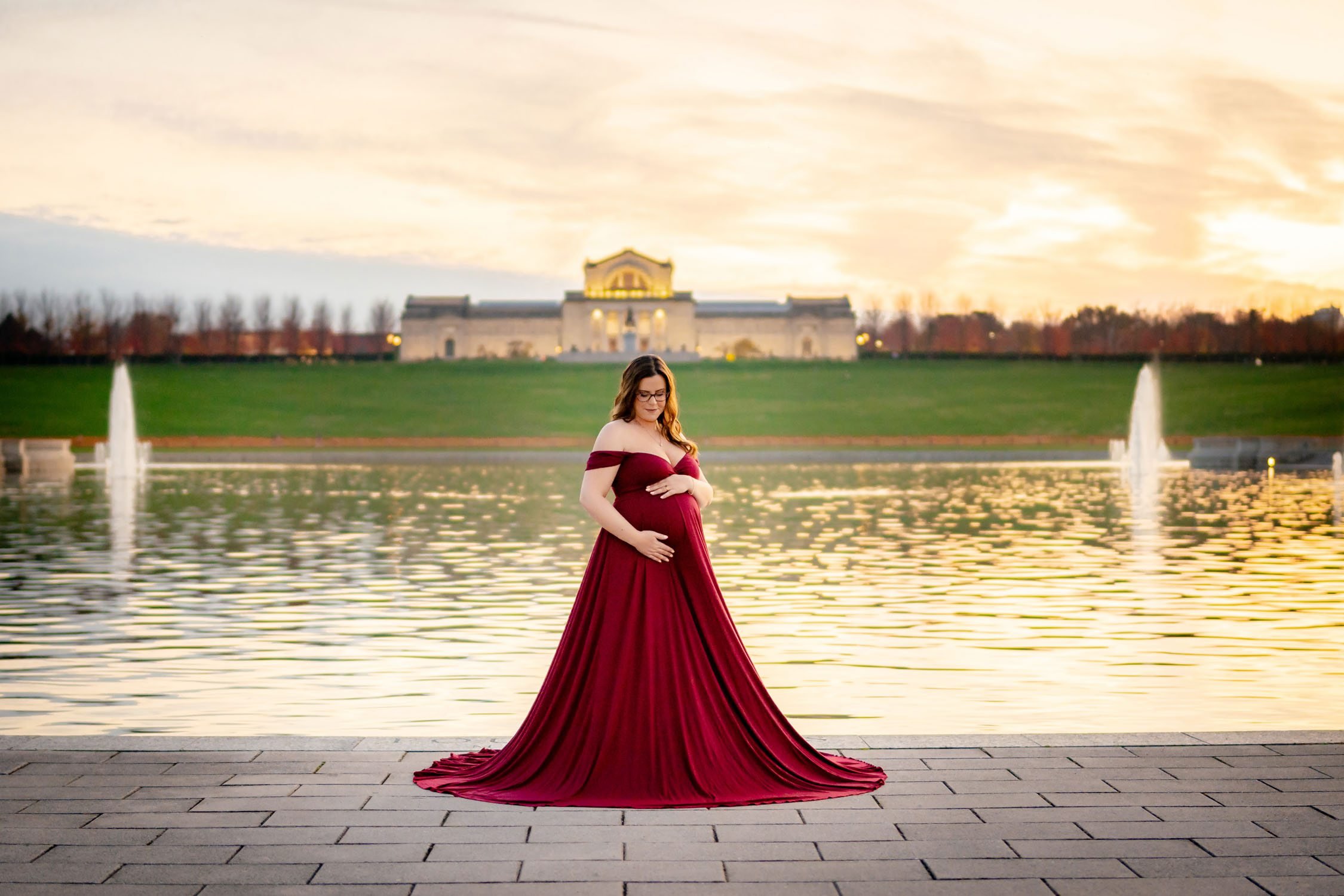 Expecting mother in a flowing red maternity gown posing beside the Grand Basin in Forest Park, St. Louis.