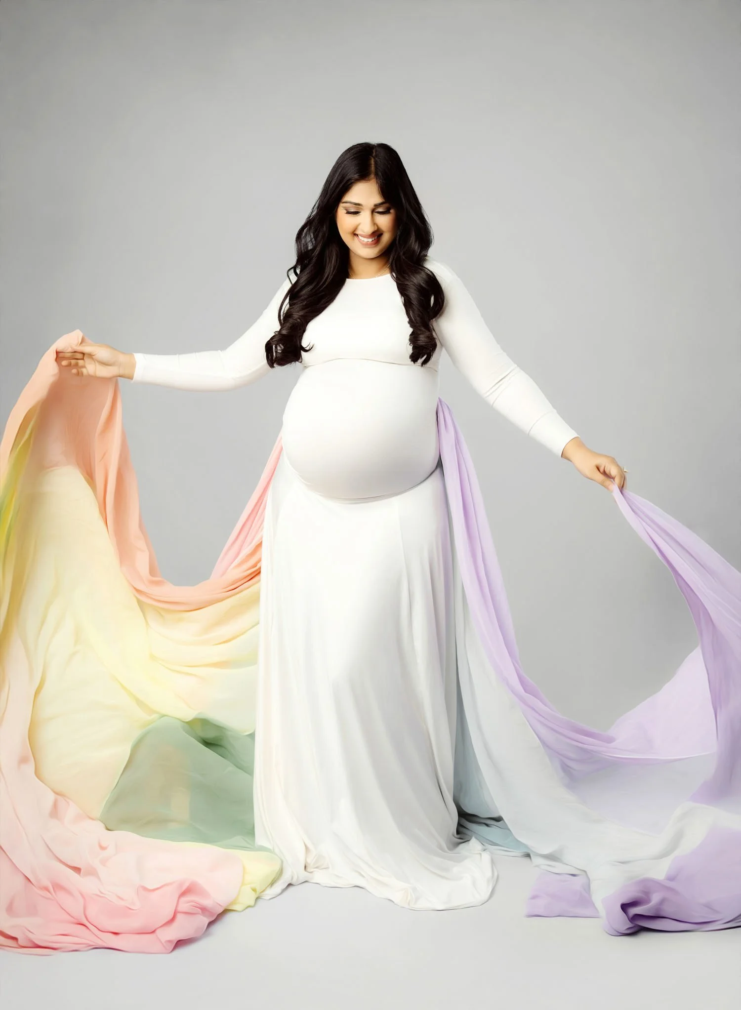 Pregnant woman in a white maternity dress holding colorful rainbow flowing fabric in the studio.