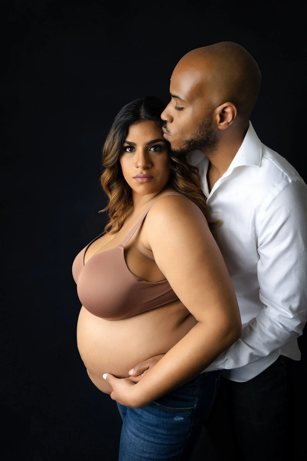 Expecting couple posing together in an intimate maternity portrait against a dark background.