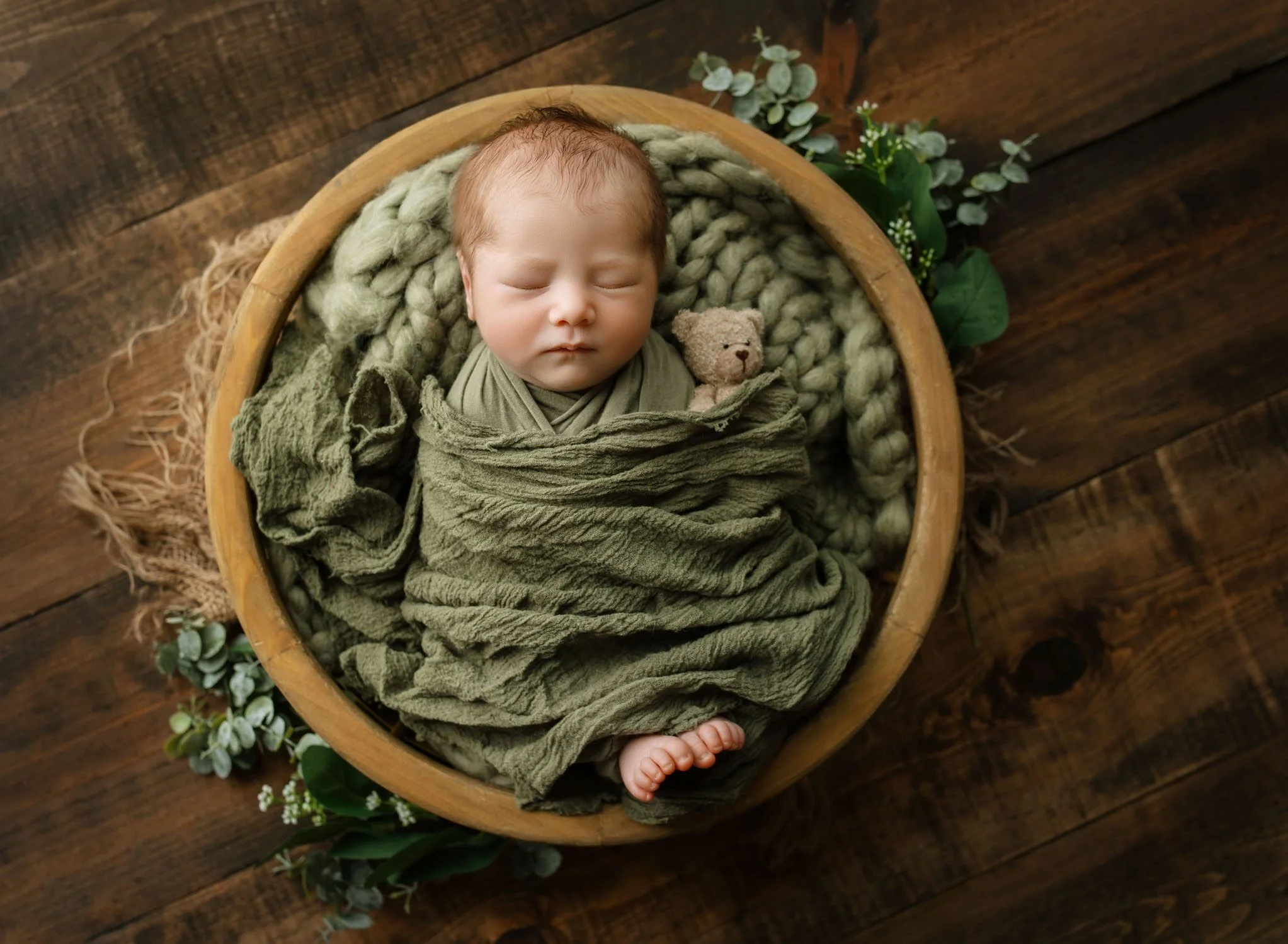 A newborn baby sleeps in a green swaddle in a wooden bowl in a studio after mom explored St. Louis baby boutiquesSt. Louis baby boutiques
