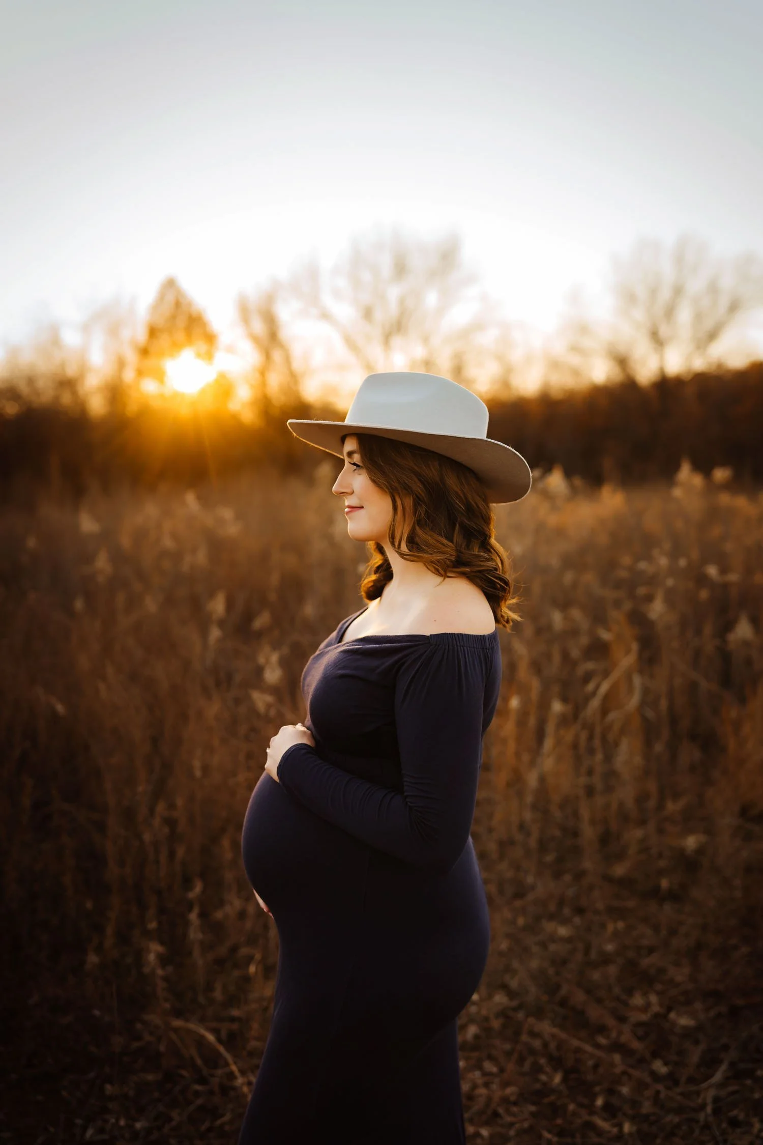 Pregnant woman in a navy gown and wide-brim hat posing in profile during an outdoor maternity portrait.