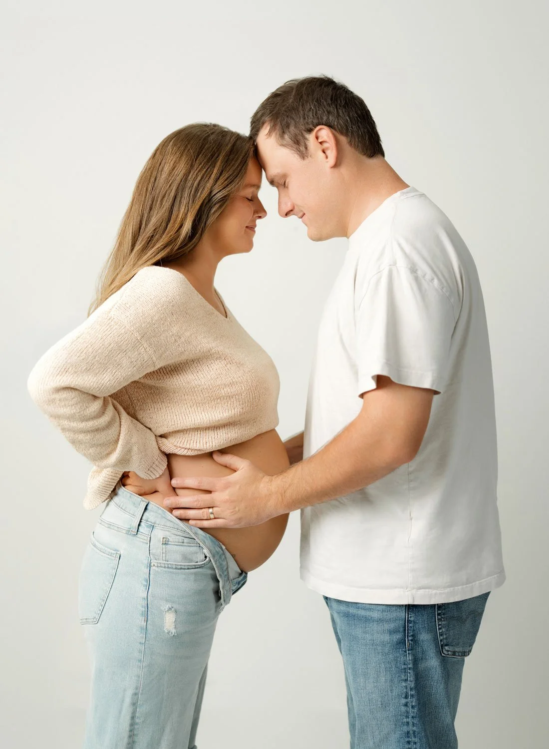 A happy pregnant couple hold the bump while touching foreheads and standing in a studio