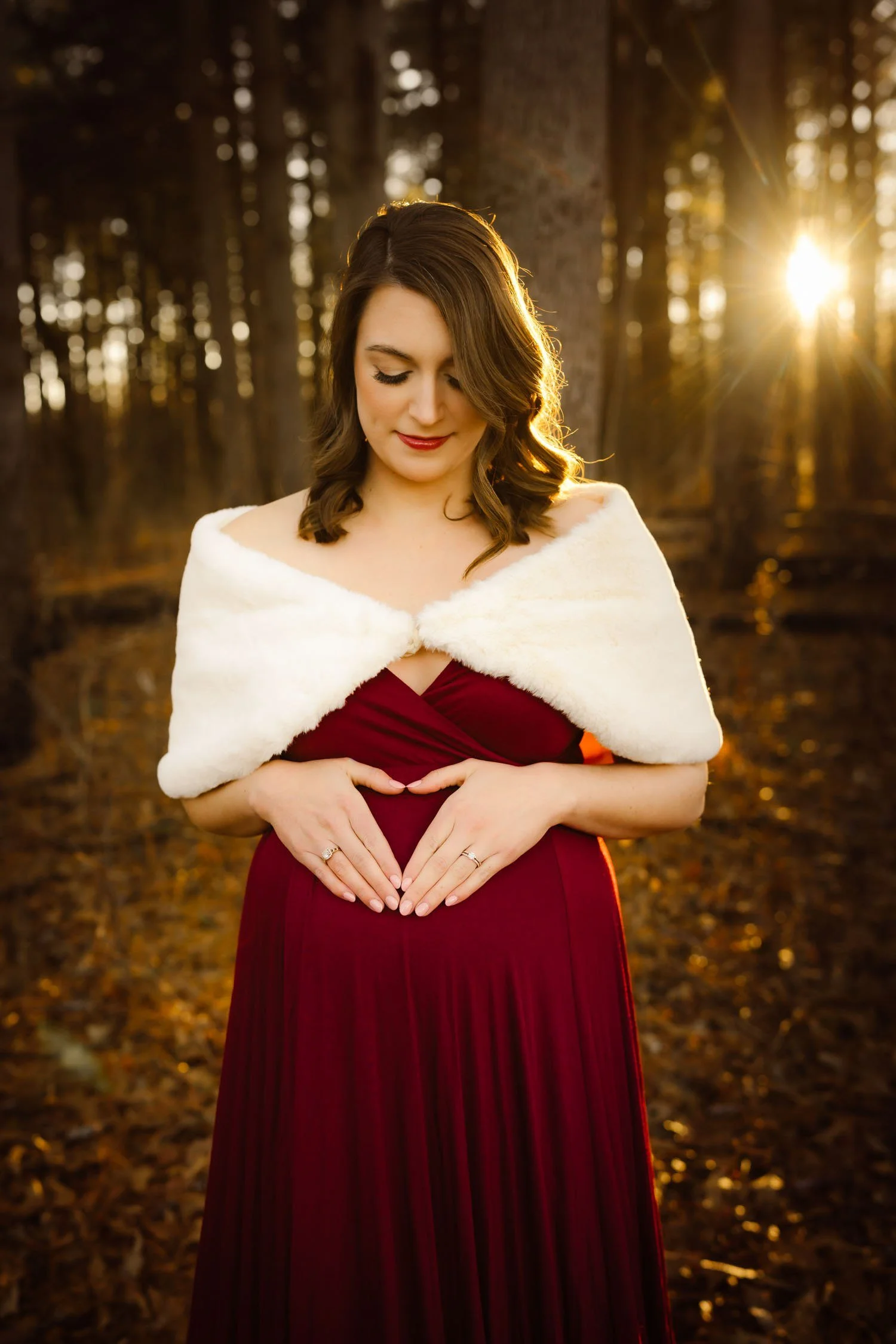 Pregnant woman in a burgundy gown and white wrap holding her baby bump during a golden-hour winter maternity session.