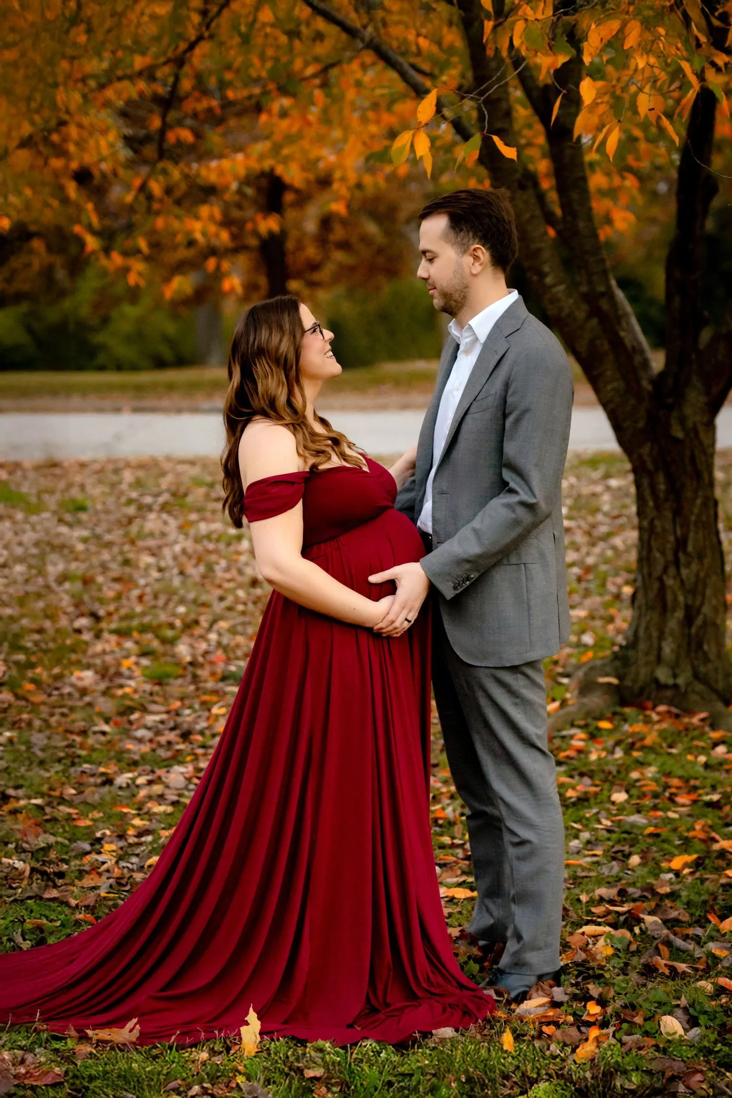 Pregnant woman in a dramatic red gown posing with her partner among fall leaves in Forest Park.