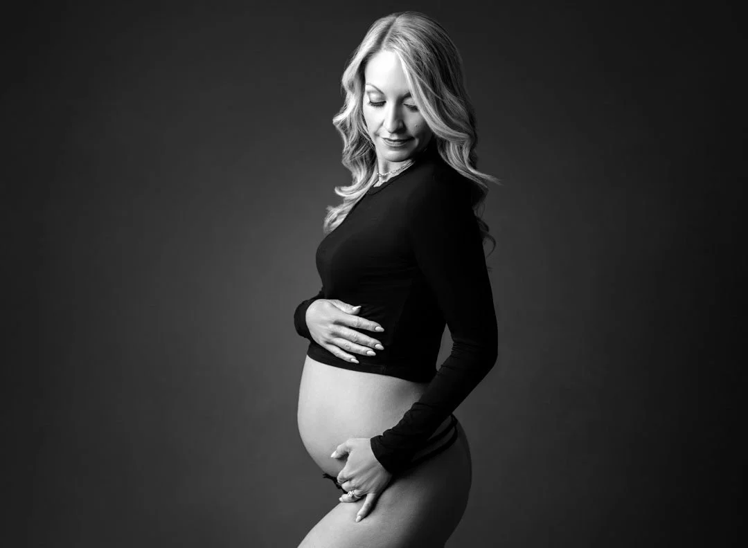 Black and white maternity portrait of a pregnant woman in a fitted black outfit holding her baby bump.
