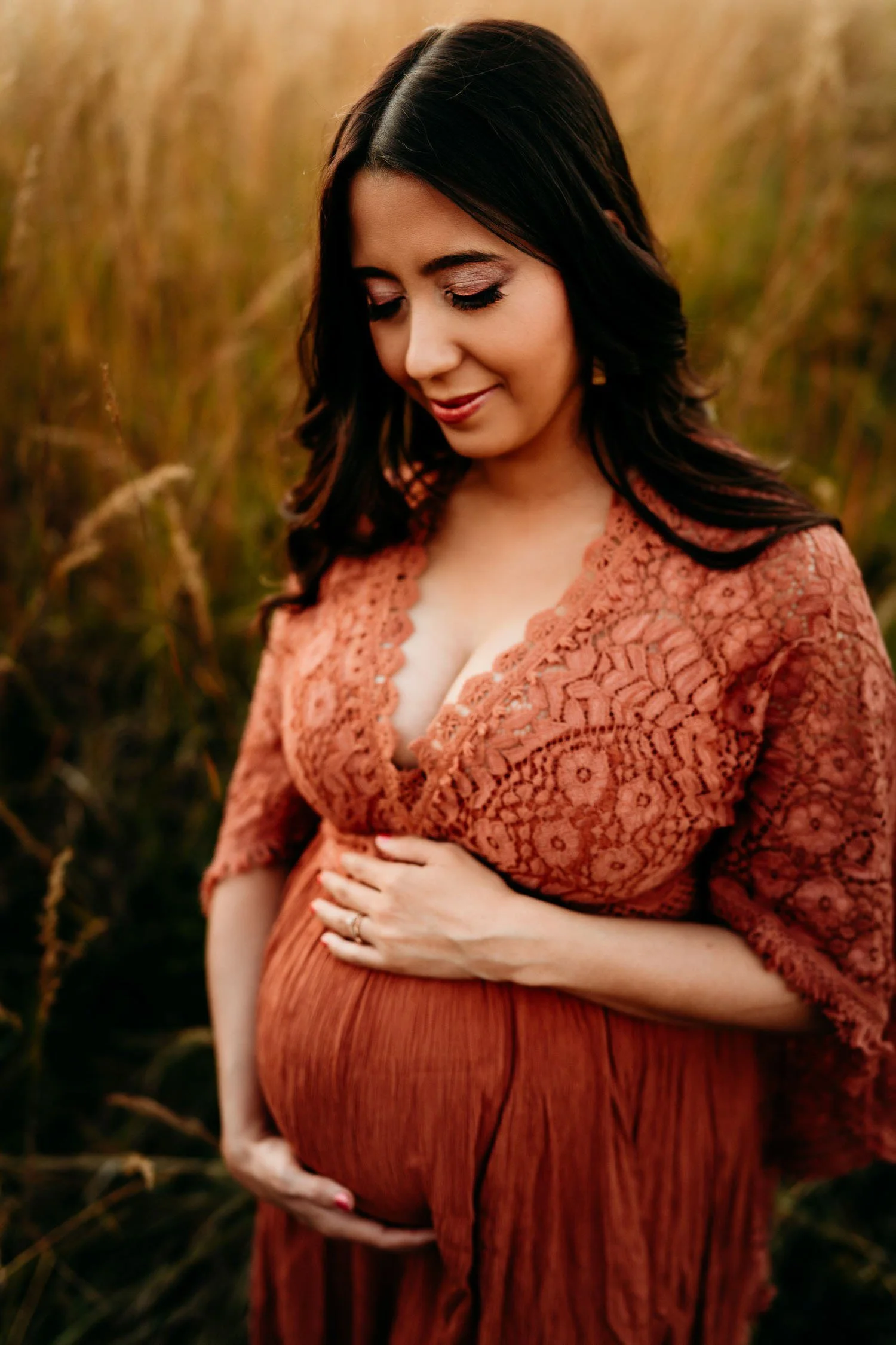 Pregnant woman in a rust lace dress cradling her baby bump in a field at sunset.