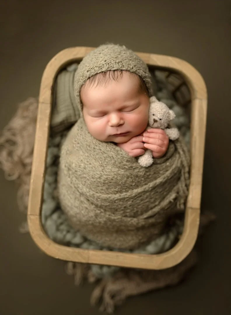 A sleeping newborn baby in a tan swaddle and matching bonnet in a wooden basket clutching a tiny teddy bear
