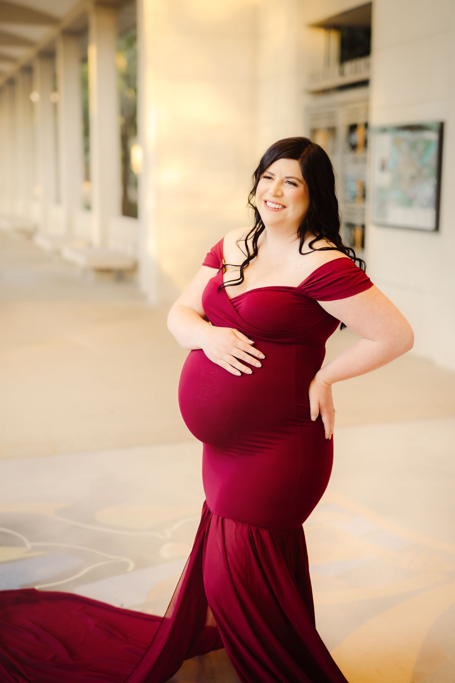 Pregnant woman in a dramatic red maternity gown posing outdoors near an architectural setting of The Muny.