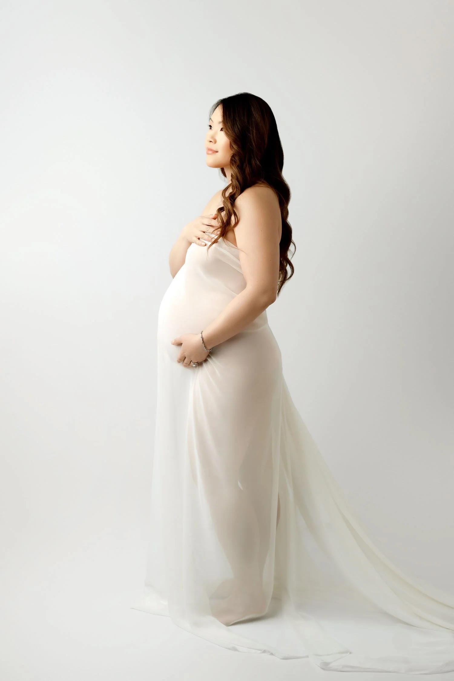 Pregnant woman in a flowing white maternity fabric posing in profile during a studio session.