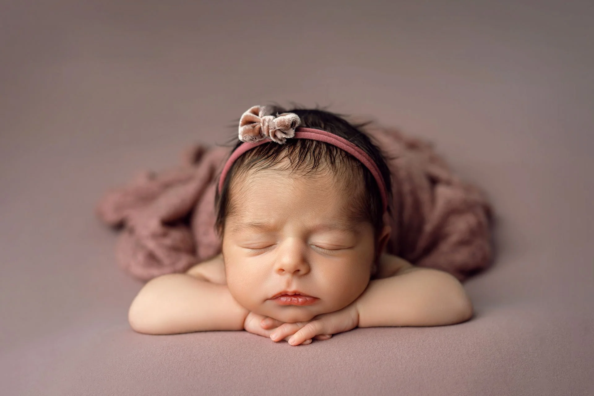 Sleeping newborn baby girl wearing a mauve bow headband, resting her chin on folded hands against a soft dusty rose backdrop