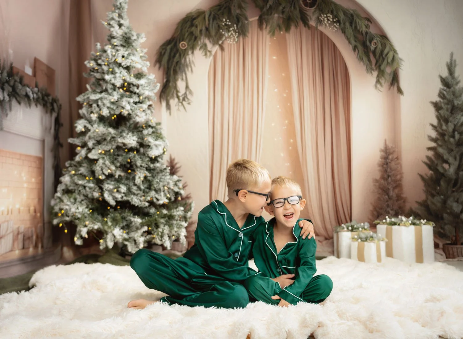 Two children in green outfits posing beside a Christmas tree on a soft neutral holiday set.