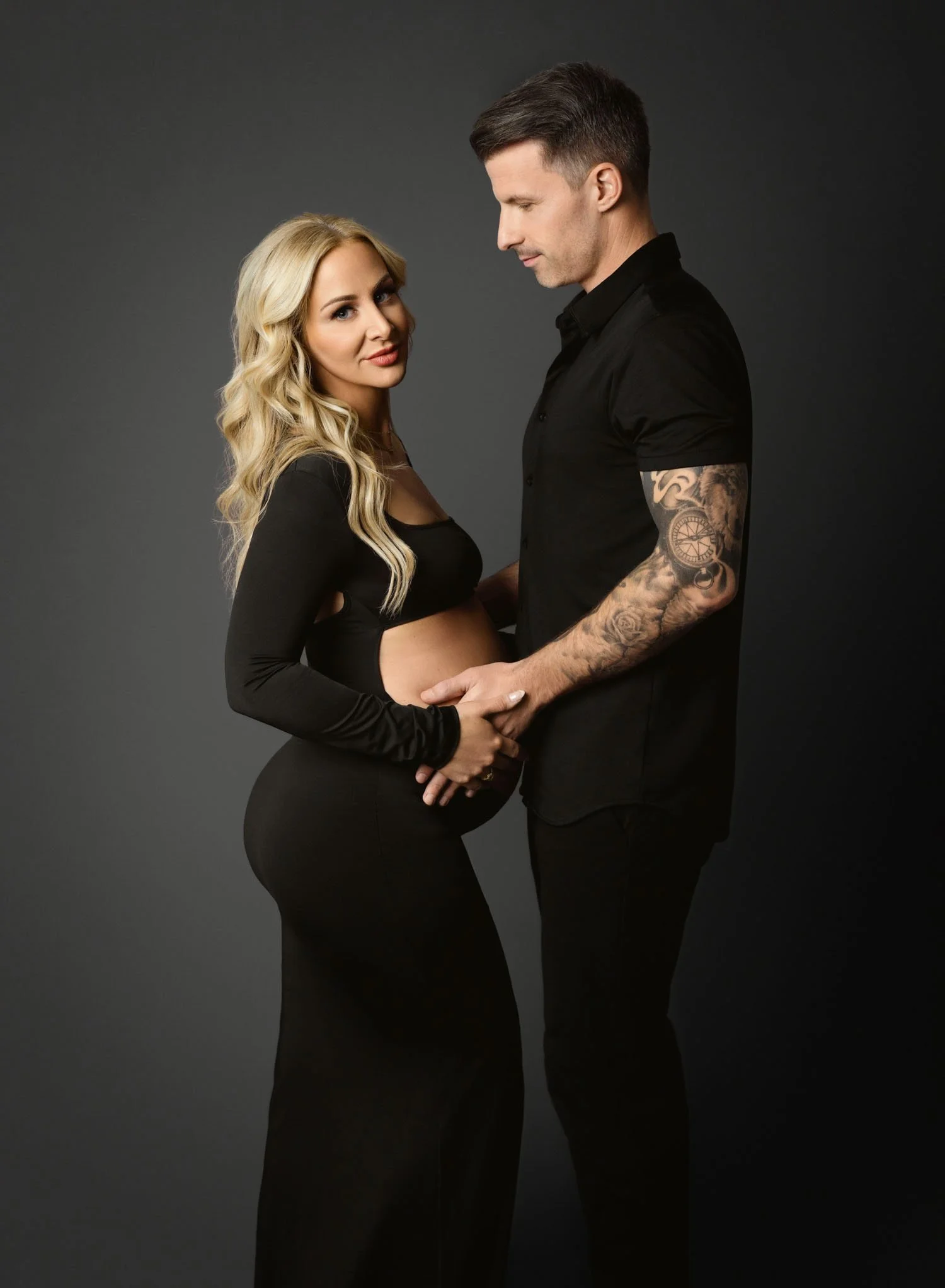 Pregnant couple holding hands over the baby bump during a black studio maternity portrait.