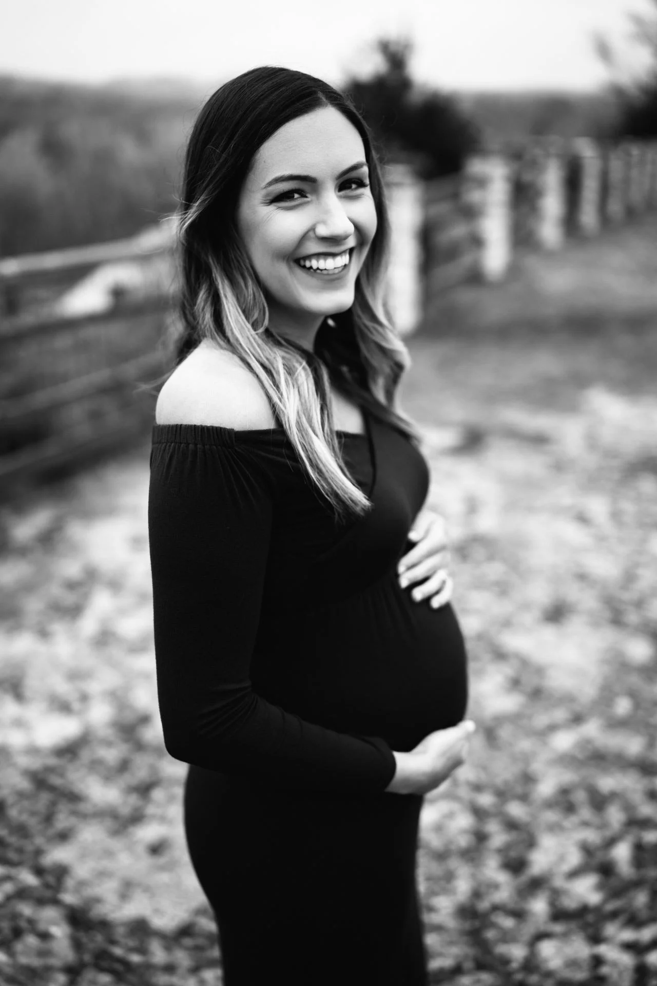 Black and white outdoor maternity portrait of a pregnant woman smiling in a fitted black dress.