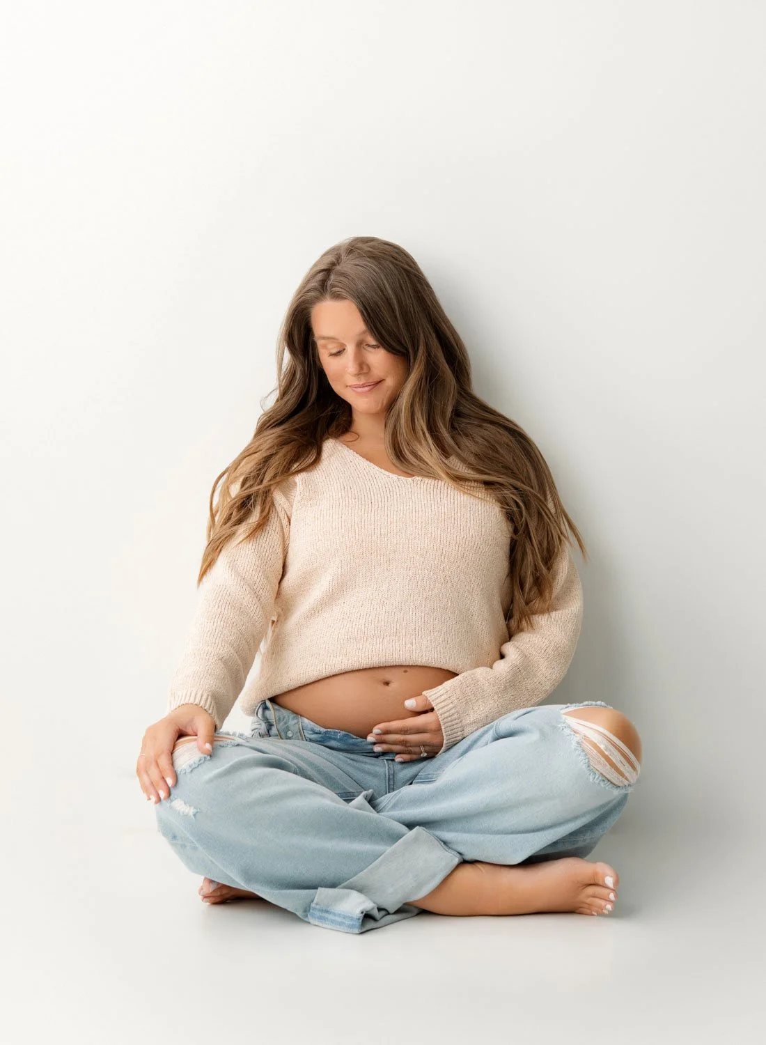 A pregnant woman sits cross legged on the floor of a studio in a tan sweater and jeans with a hand under her bump after meeting midwives in St. Louis