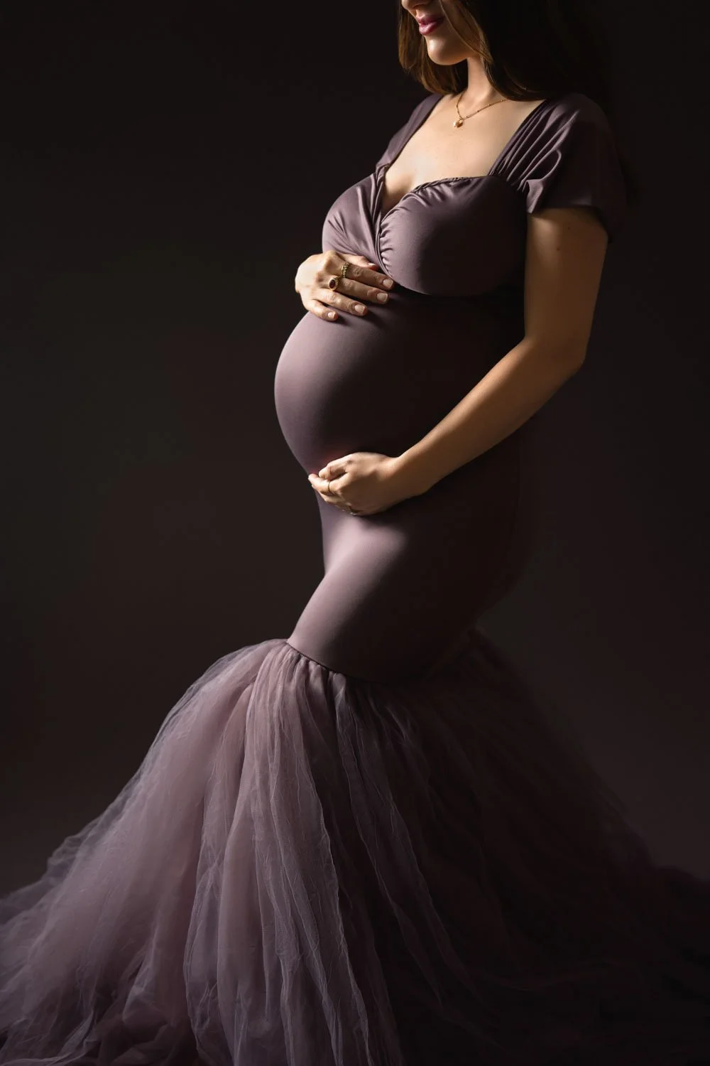 Close maternity portrait of an expecting mother in a fitted mauve gown holding her baby bump.