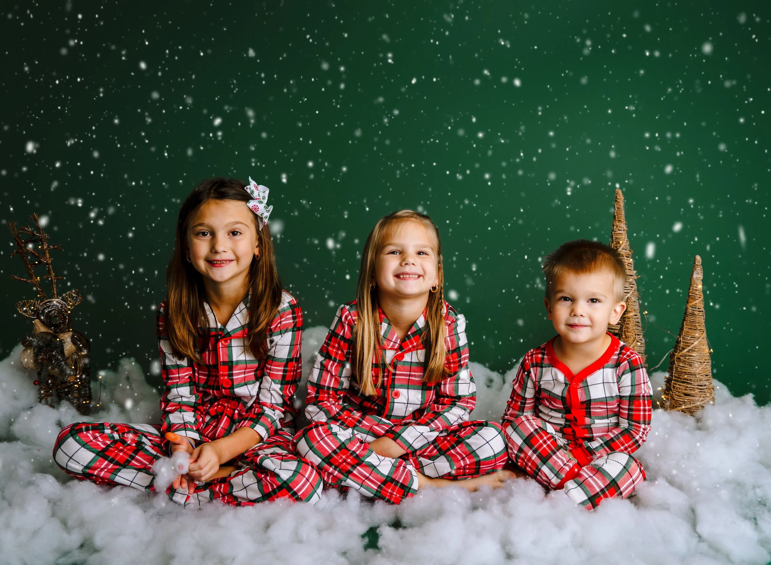 Three children in matching Christmas pajamas posing on a green holiday studio set.