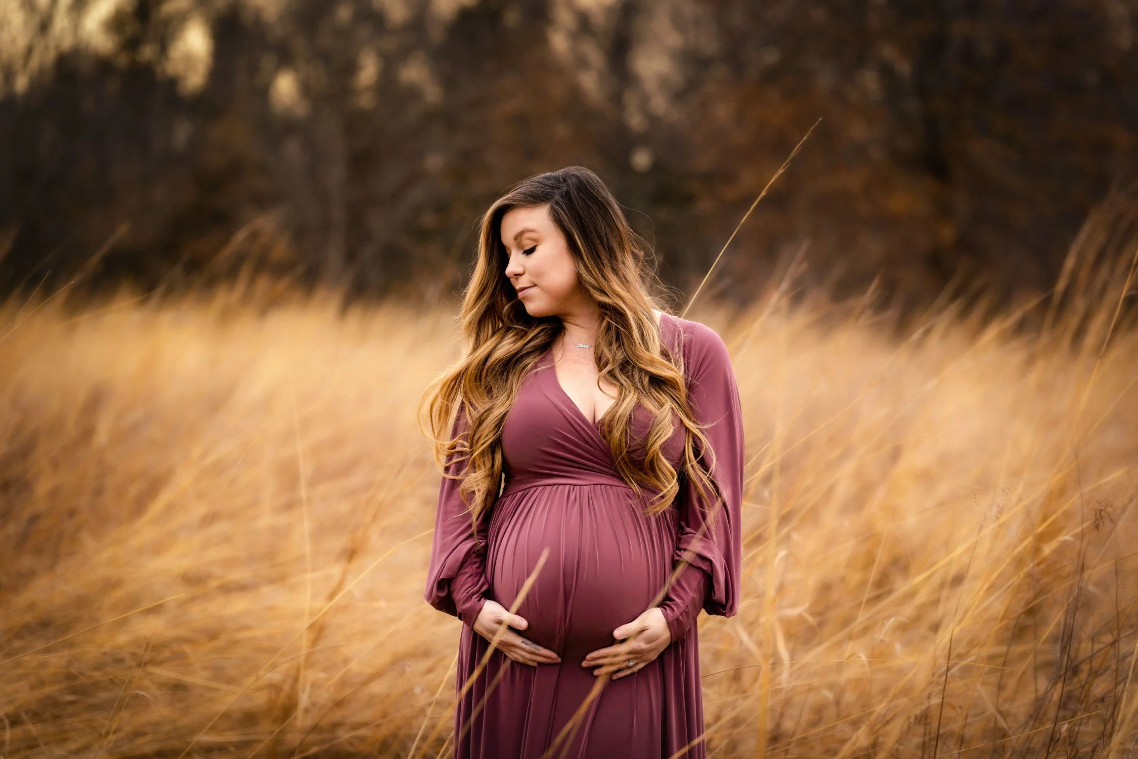 Pregnant woman in a mauve dress standing in a field of tall golden grass.