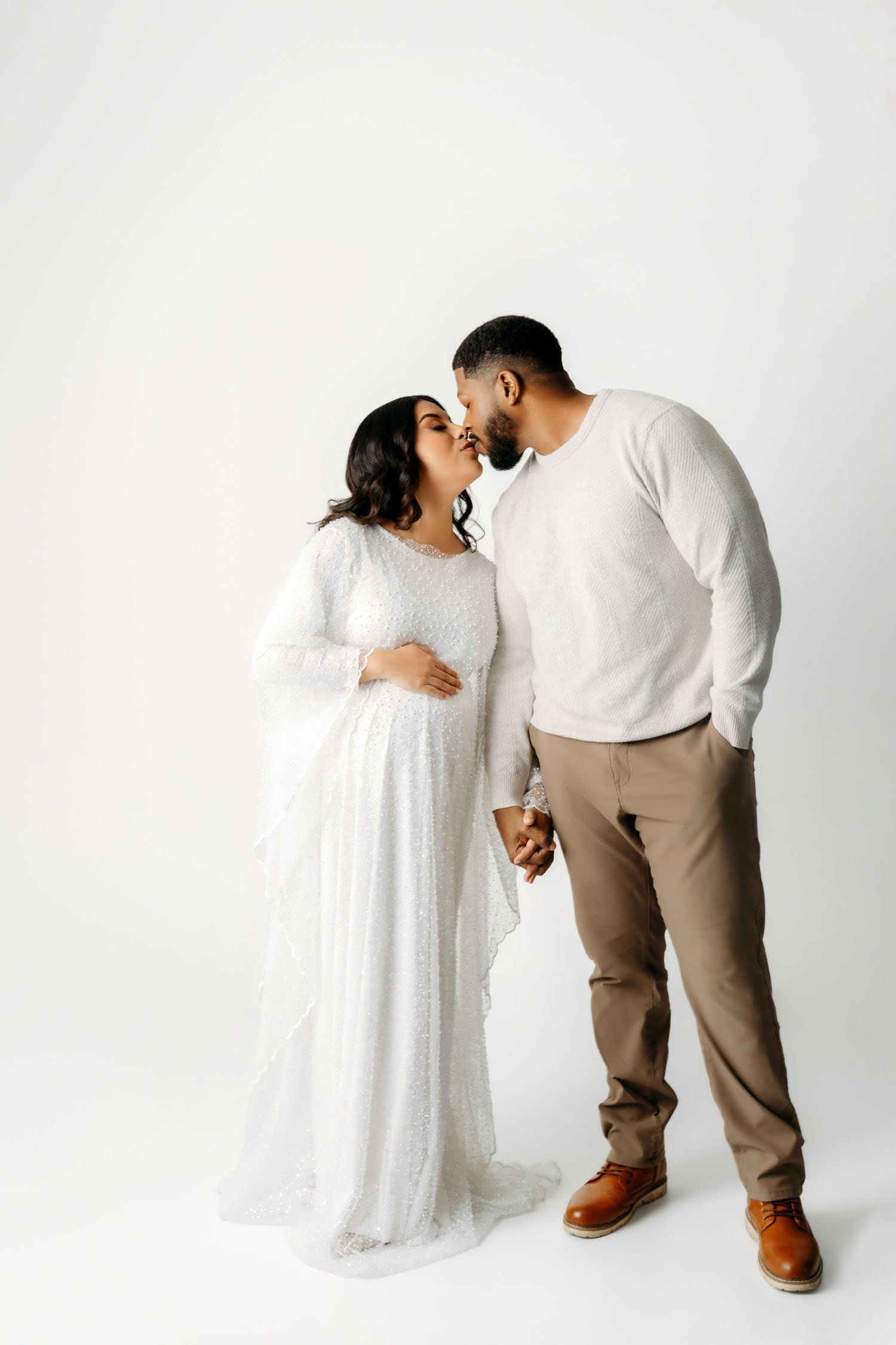 Expecting couple sharing a kiss while holding hands during a bright studio maternity portrait.