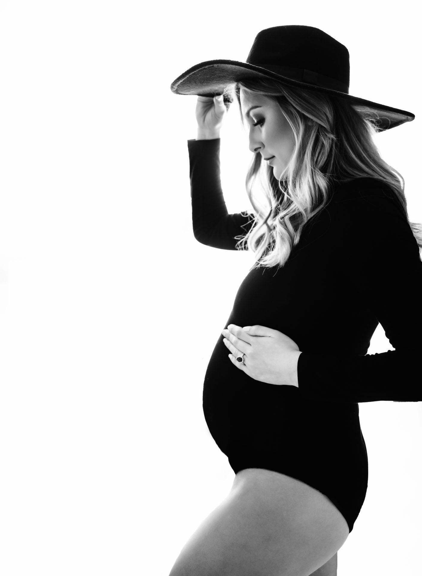 Black and white maternity portrait of a pregnant woman in a wide-brim hat and fitted black outfit.