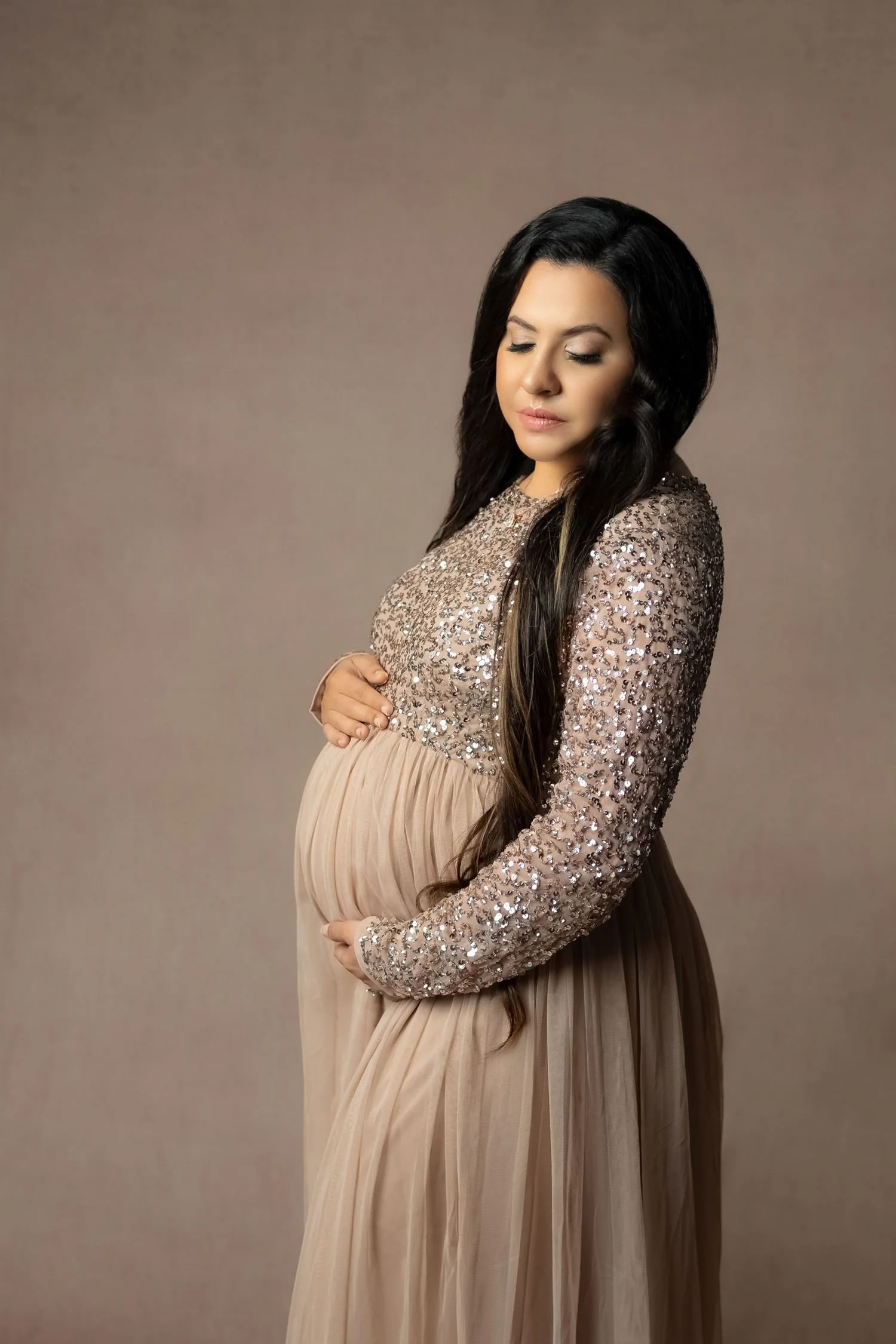 Expecting mother in a sparkling neutral gown cradling her baby bump.