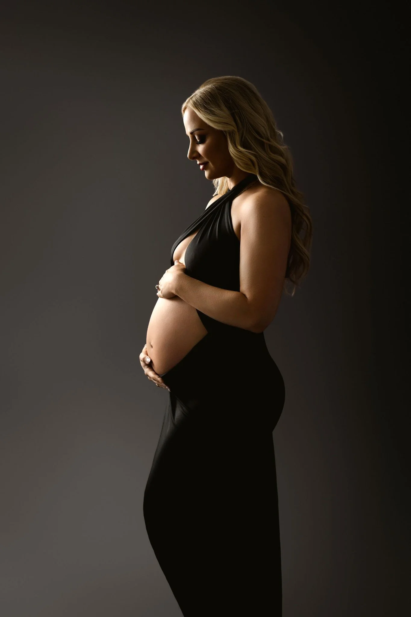 A smiling mother to be holds her hands on her bump in a black open front top in a studio after visiting birth center in st. louis