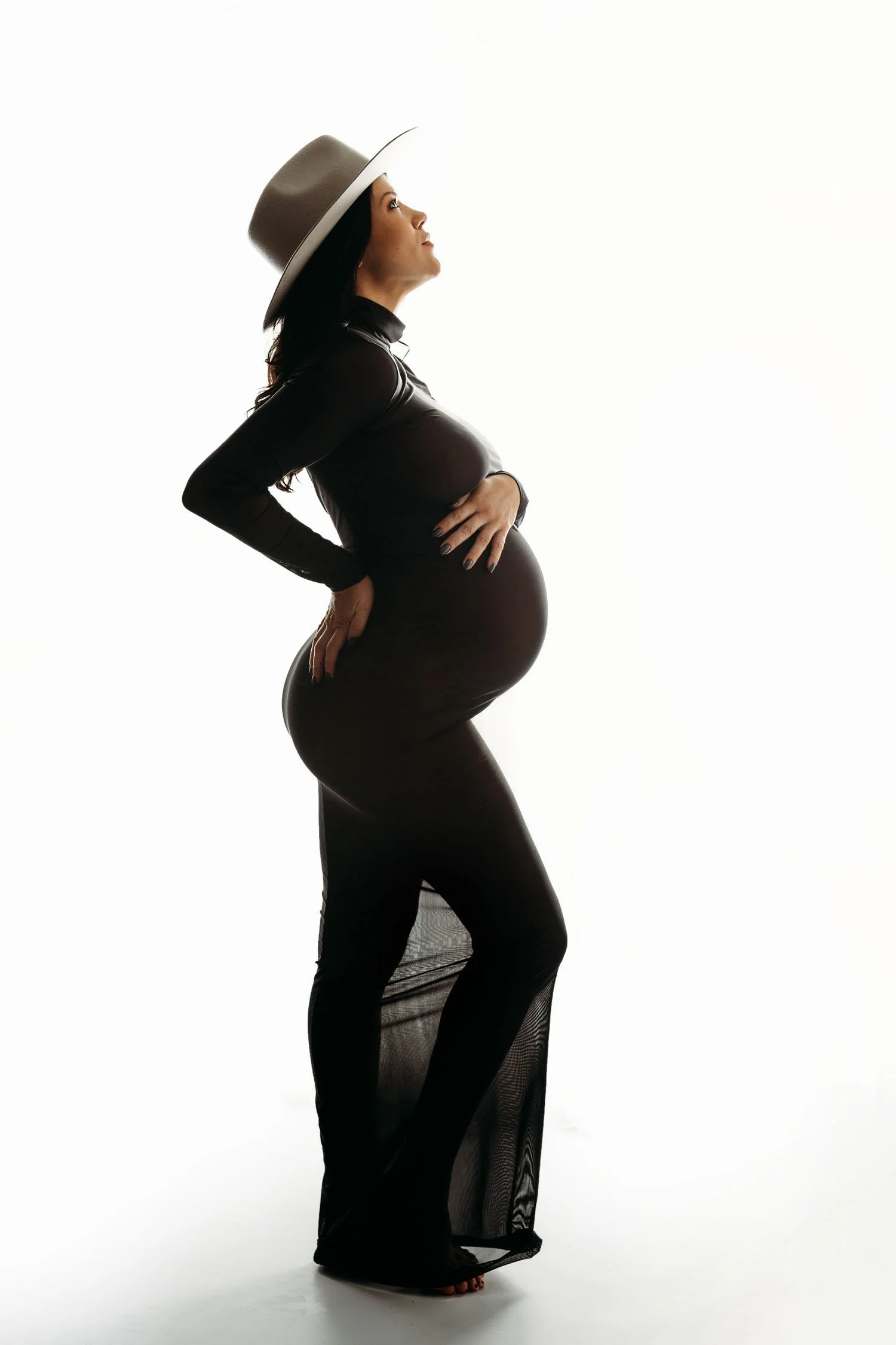 Pregnant woman in a fitted black gown and wide-brim hat posing in profile during a St. Louis pregnancy photography session.