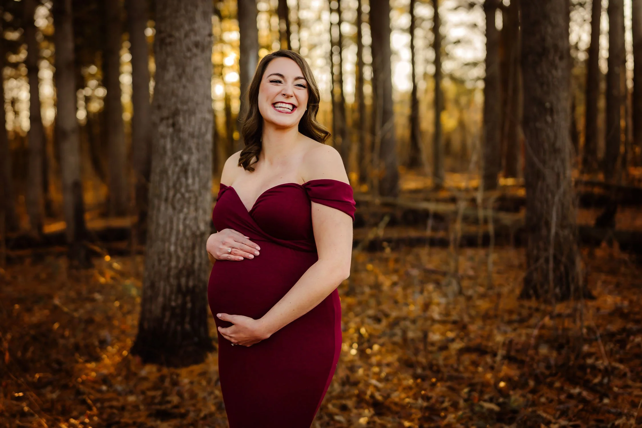 Expecting mother in a burgundy gown smiling in a wooded setting during a St. Louis maternity photography session.