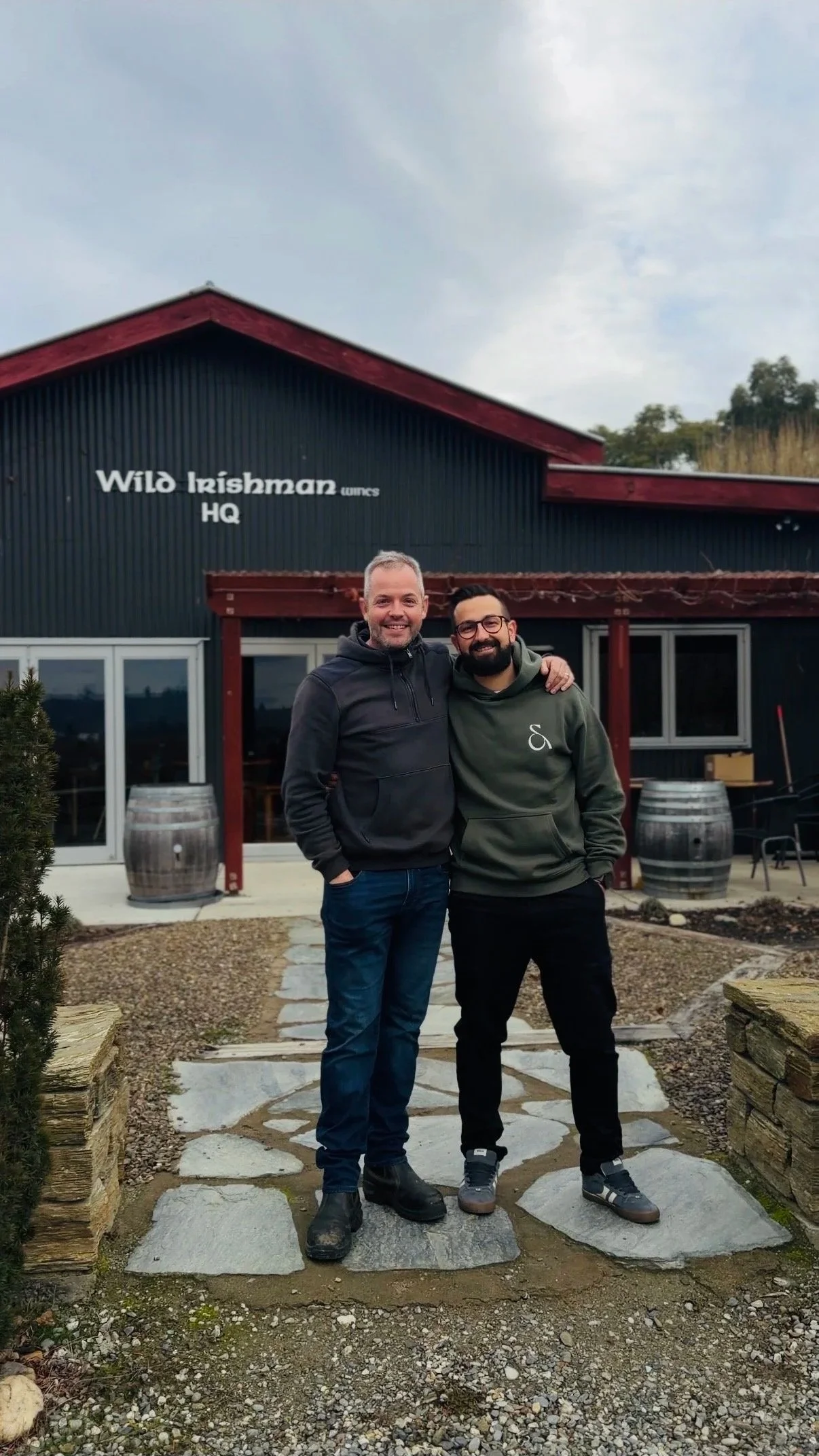 Two men standing outside a restaurant called 'Wild Irishman' with their arms around each other, smiling, in front of a black building with a red roof, and weathered wine barrels. One is wearing a black hoodie and jeans, the other a green hoodie and black pants.