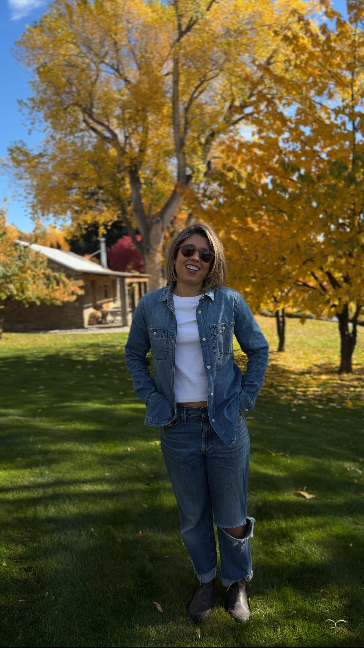 A woman with short hair and sunglasses smiling and posing outdoors in front of trees with autumn-colored leaves. She's wearing a denim jacket, white shirt, ripped jeans, and boots.
