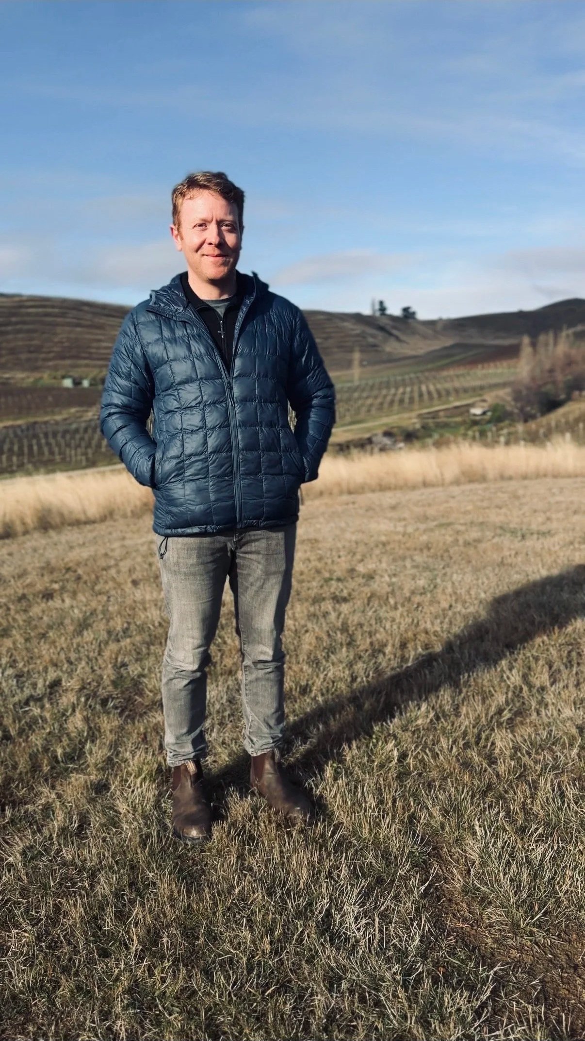 A man standing outdoors in front of a hilly landscape with vineyards, wearing a blue quilted jacket, gray pants, and brown shoes, with a blue sky overhead.