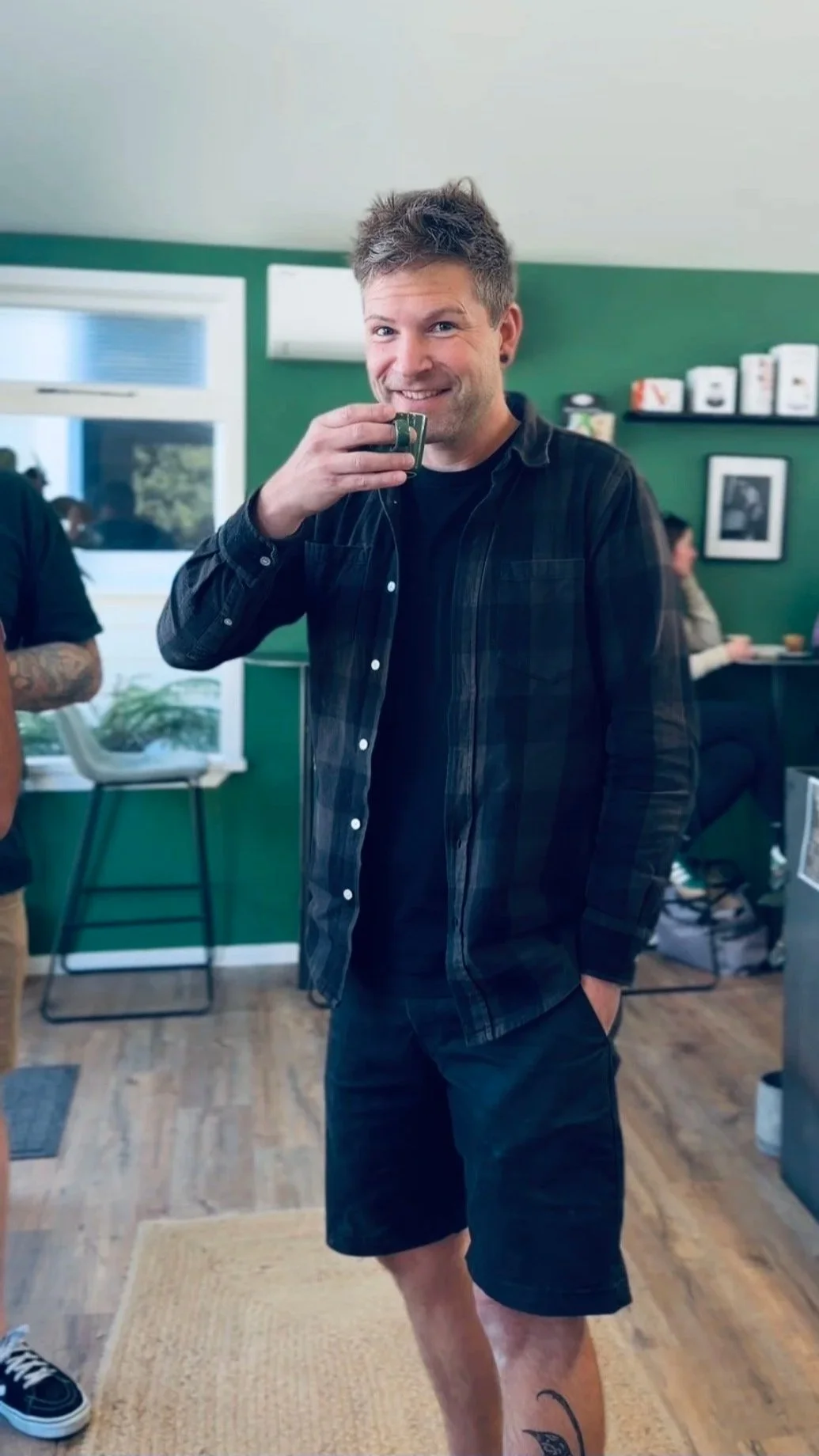 A young man with short brown hair and earrings smiling while drinking coffee in a cozy room with green walls, bookshelf, and framed photo, with people in the background.