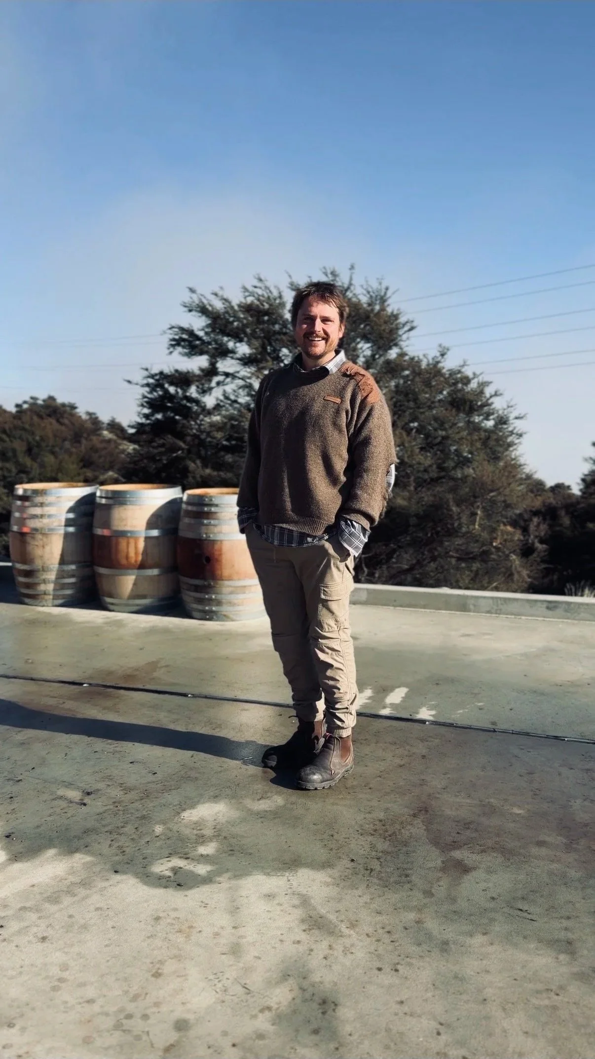 A man standing outdoors on a rooftop or parking structure with barrels in the background and trees, wearing a brown sweater, plaid shirt, khaki cargo pants, and boots, smiling at the camera.