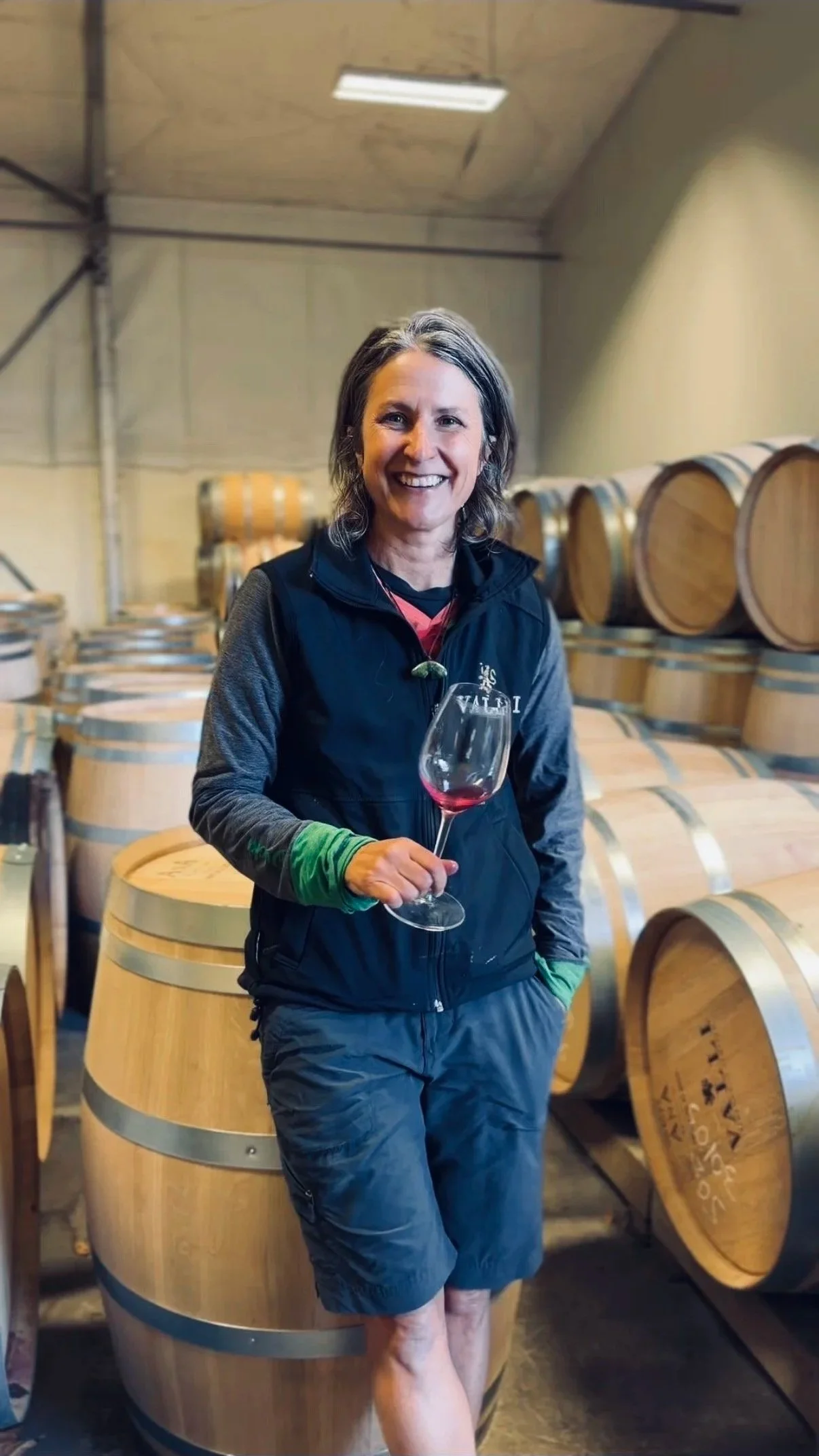 A woman with shoulder-length gray hair smiling and holding a glass of red wine, standing among wine barrels in a winery or cellar.