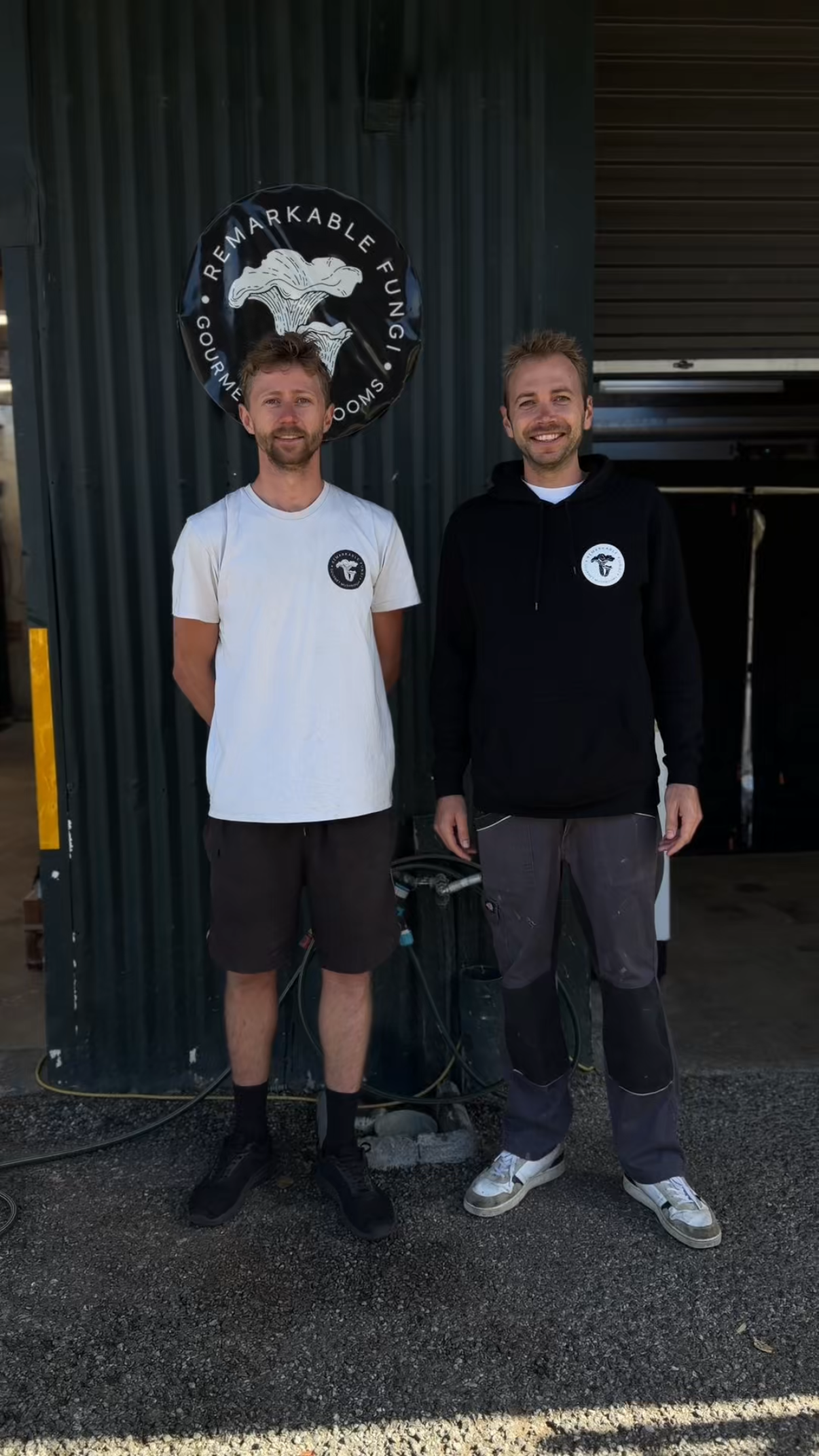 Two men standing outside in front of a dark green corrugated metal wall with a round black sign that says 'Remarkable Fungi' and features a white mushroom illustration. The man on the left is wearing a white t-shirt with a black logo, shorts, and sneakers, and the man on the right is wearing a black hoodie with a logo, cargo pants, and sneakers. Both are smiling.