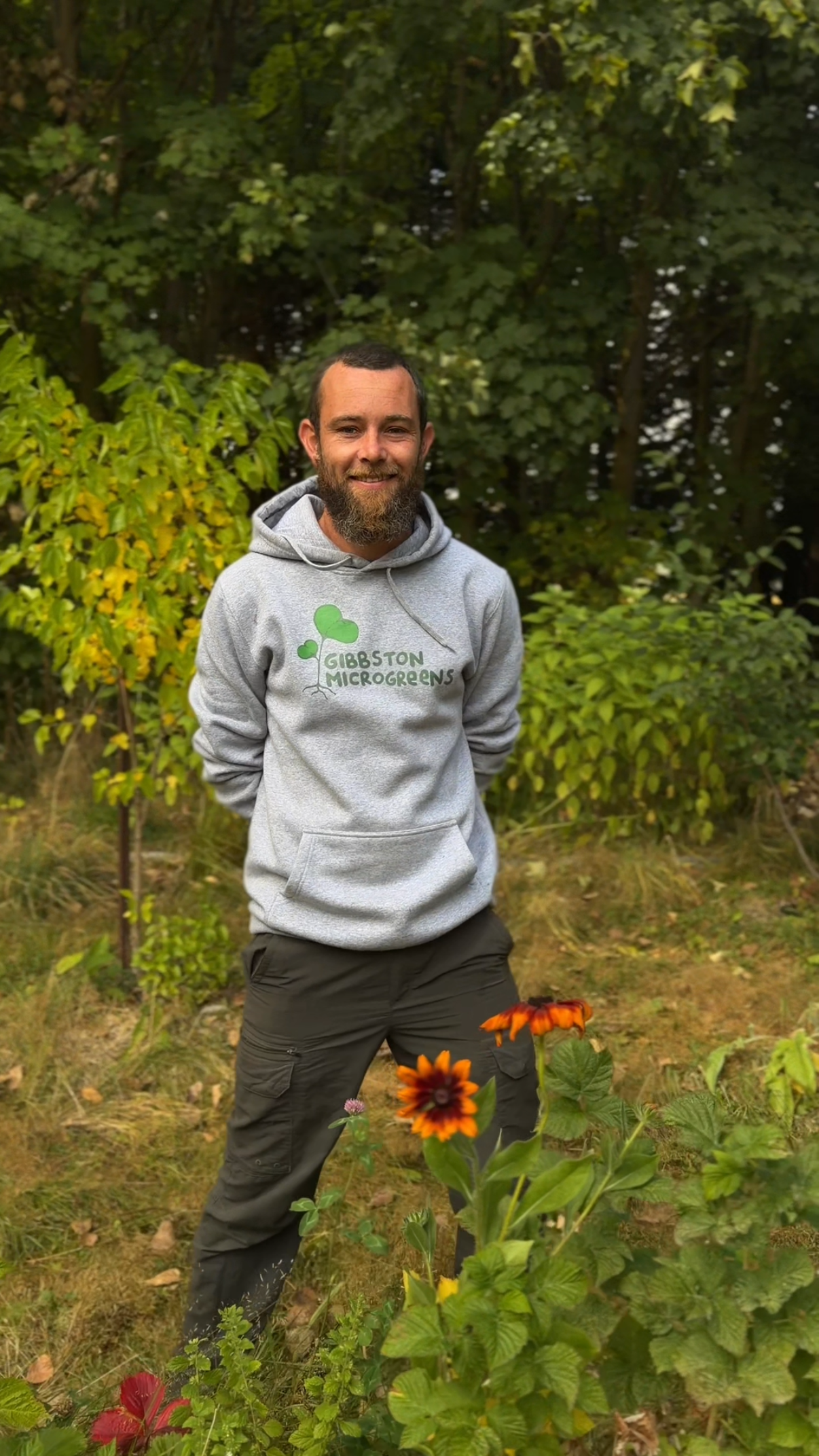 A man standing outdoors in front of green trees and bushes, wearing a gray hoodie with 'GIBBSTON MICROGREENS' and a green logo, dark pants, and smiling.