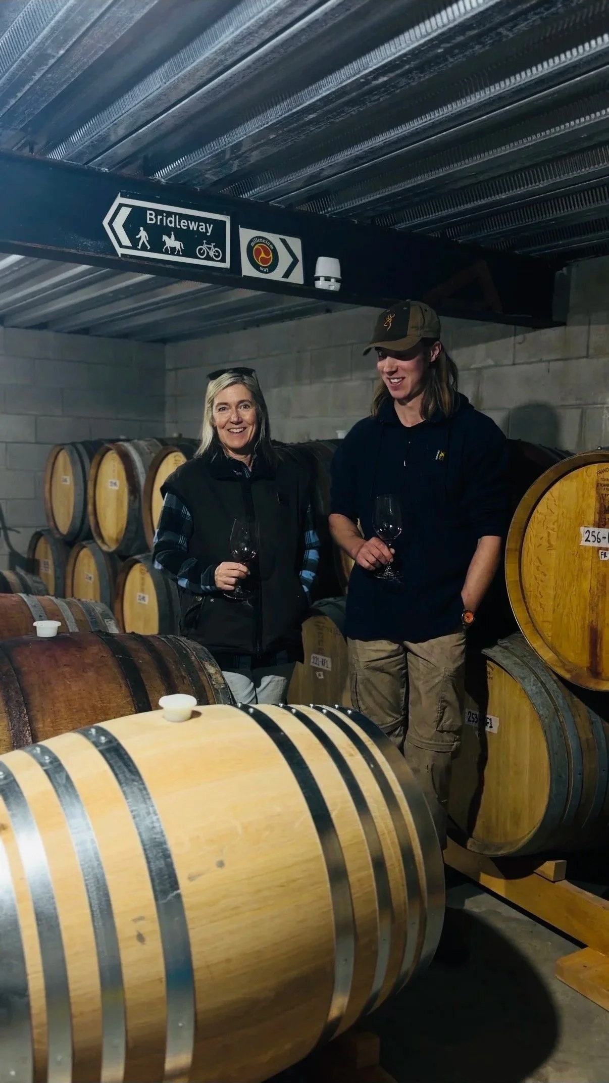 Two women holding wine glasses inside a wine cellar, standing among large wooden wine barrels, with a sign overhead guiding to a bridleway for walking, horses, and cycling.