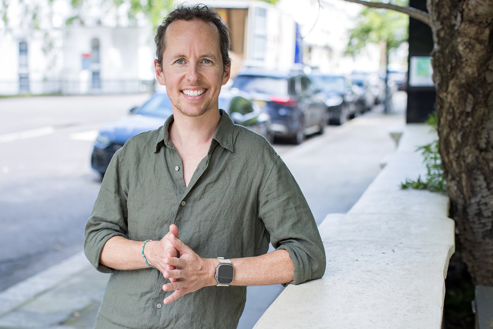 A smiling man with short dark hair, wearing a green button-up shirt and a smartwatch, standing outdoors on a sunny day beside a white stone wall, with parked cars and trees in the background.