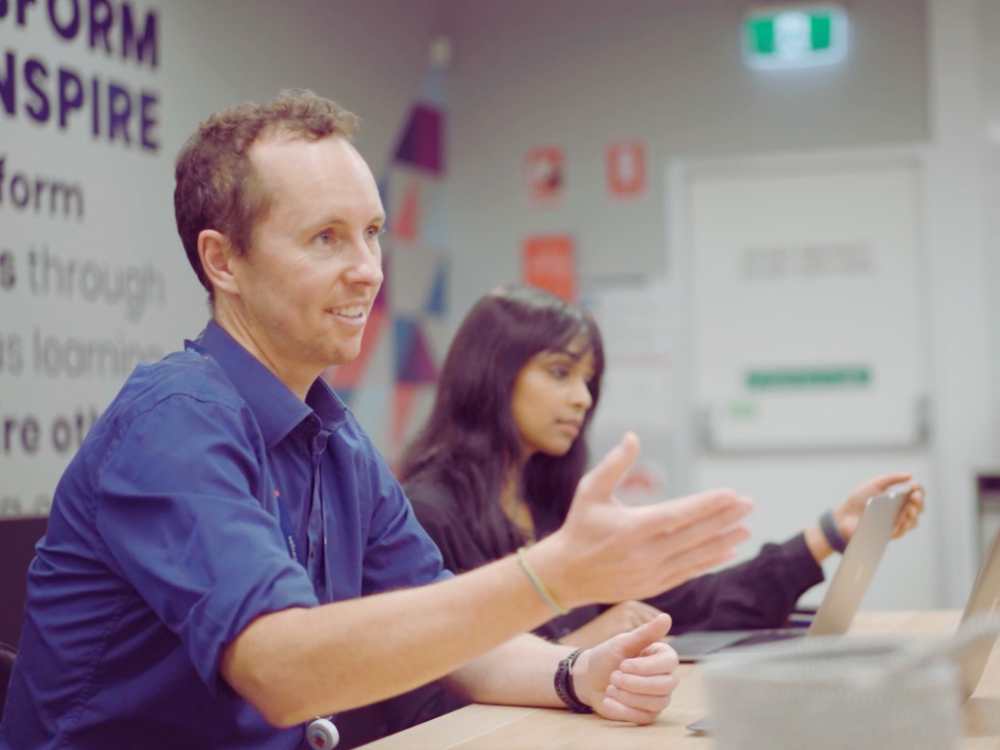 Man and woman sitting at a table with laptops, engaging in a discussion in a classroom or workshop setting.