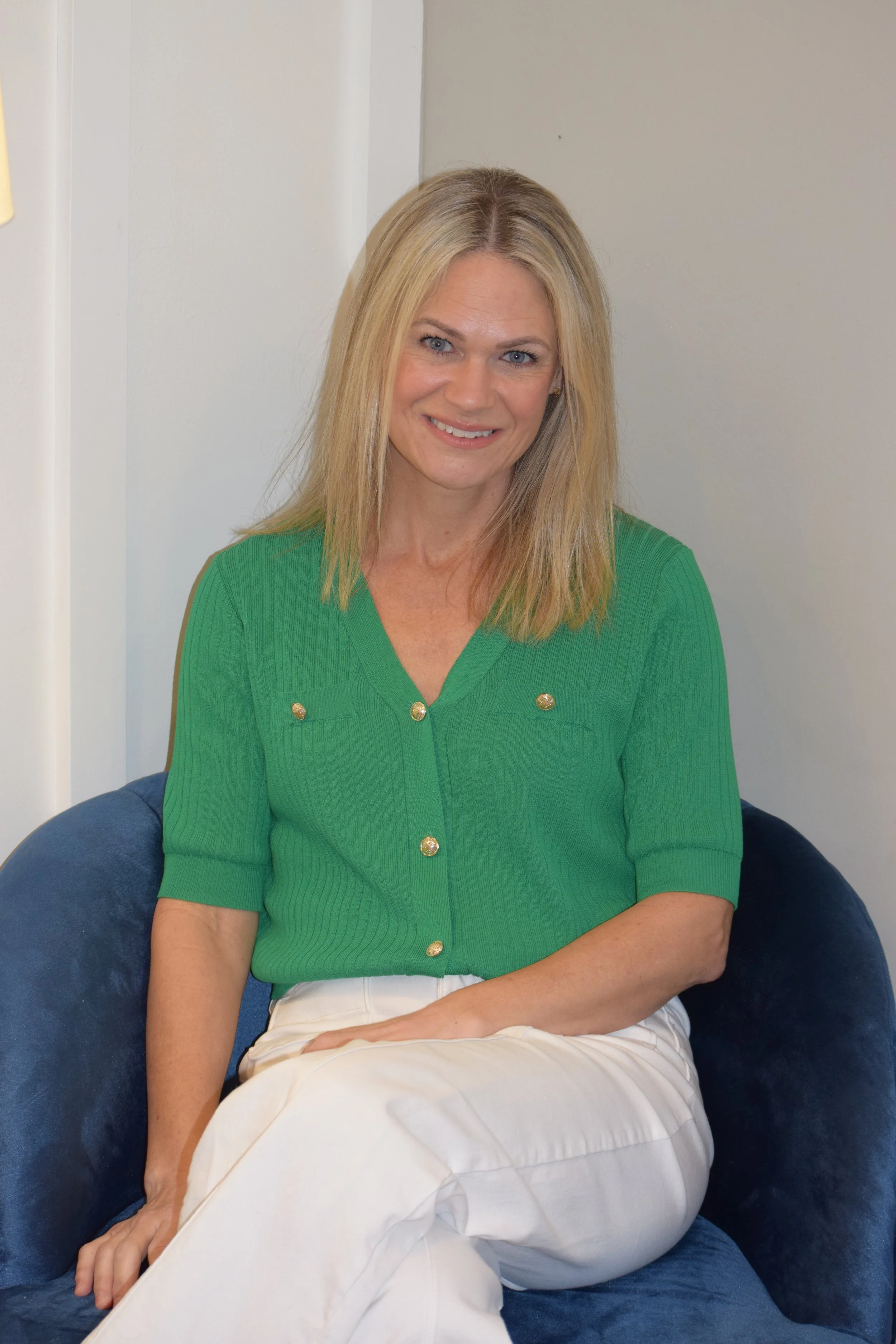 Smiling woman with blonde hair wearing a green top and white pants, sitting on a blue chair in a room with plain walls.