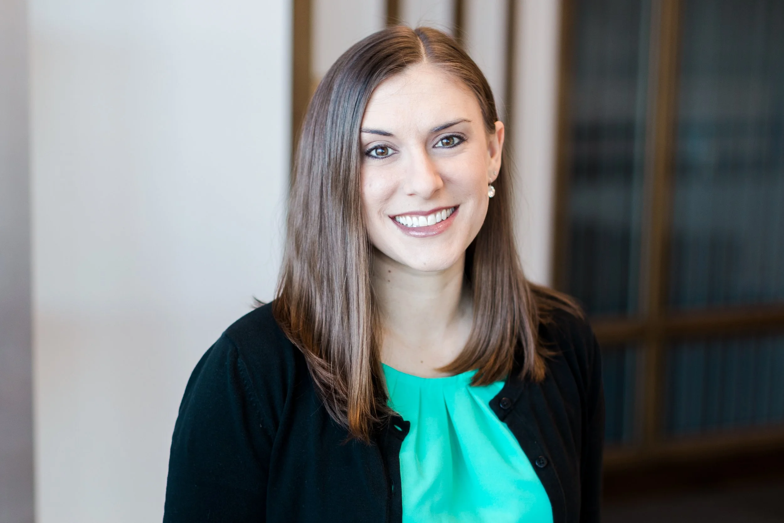 A woman with shoulder-length brown hair, wearing a black cardigan over a turquoise blouse, smiling in an indoor setting.
