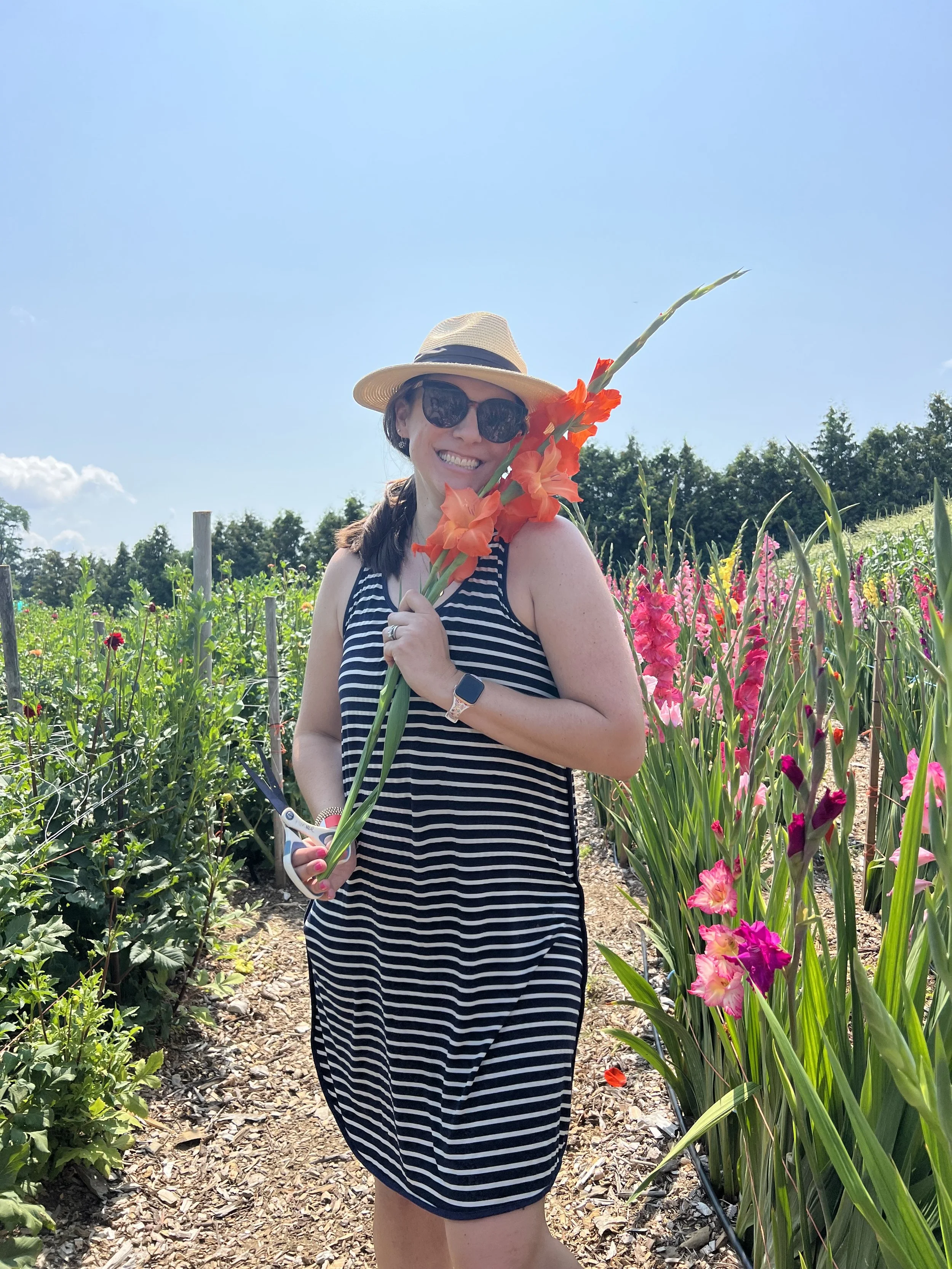 Woman in a striped dress and sunhat holding a large orange flower, standing in a colorful flower garden under a clear blue sky.