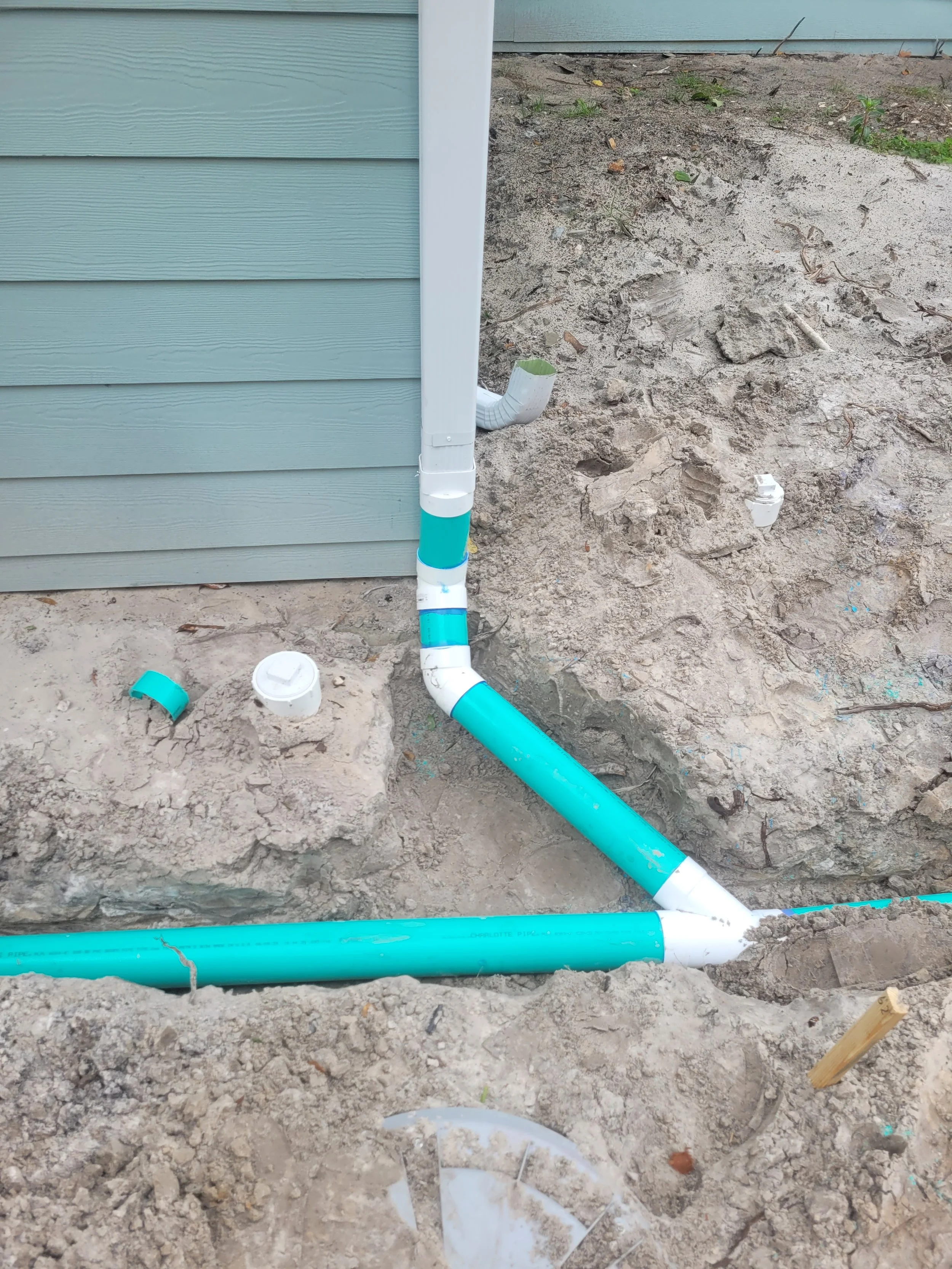 Underground plumbing pipes and fittings installed outside a house with siding, including a large teal pipe and white fittings, some of which have blue tape, surrounded by dirt and construction debris.