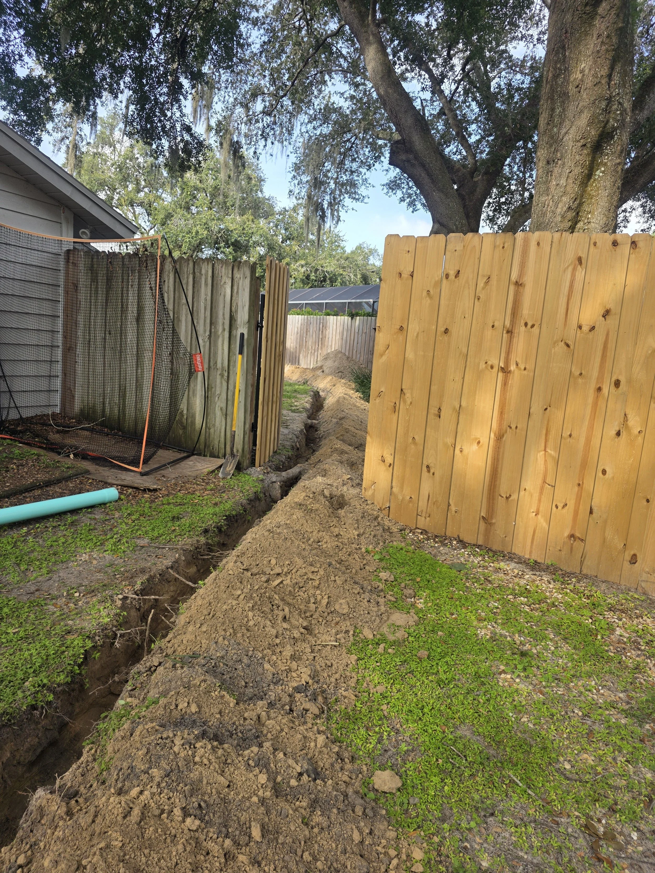 A backyard with a trench dug for fence installation or repair, a wooden fence on the right side, a wire mesh fence on the left, a large tree in the background, and construction tools such as a shovel and a level nearby.