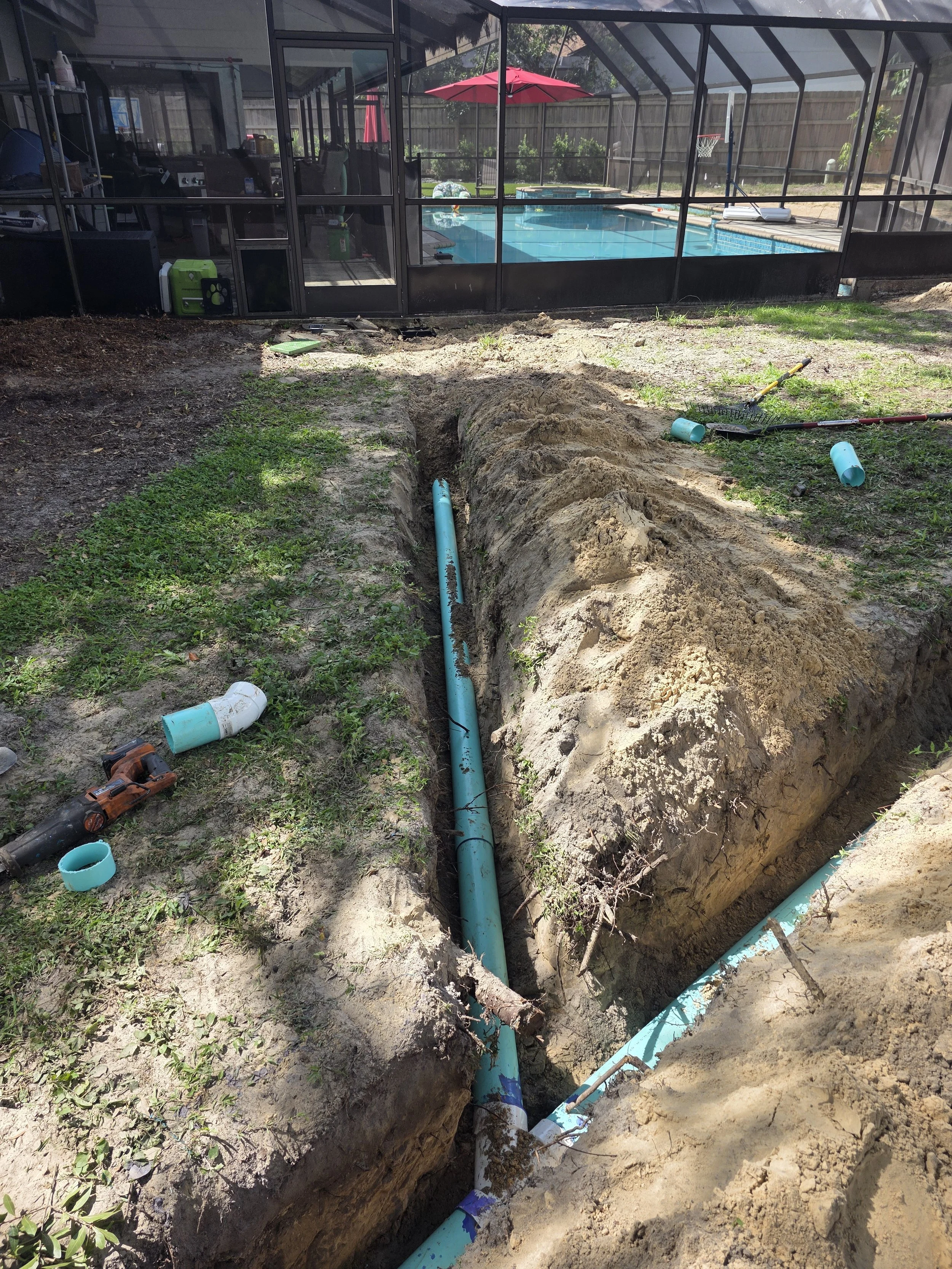 Underground plumbing work with blue pipes in a trench in a backyard, near a screened-in pool enclosure and a backyard with a basketball hoop.