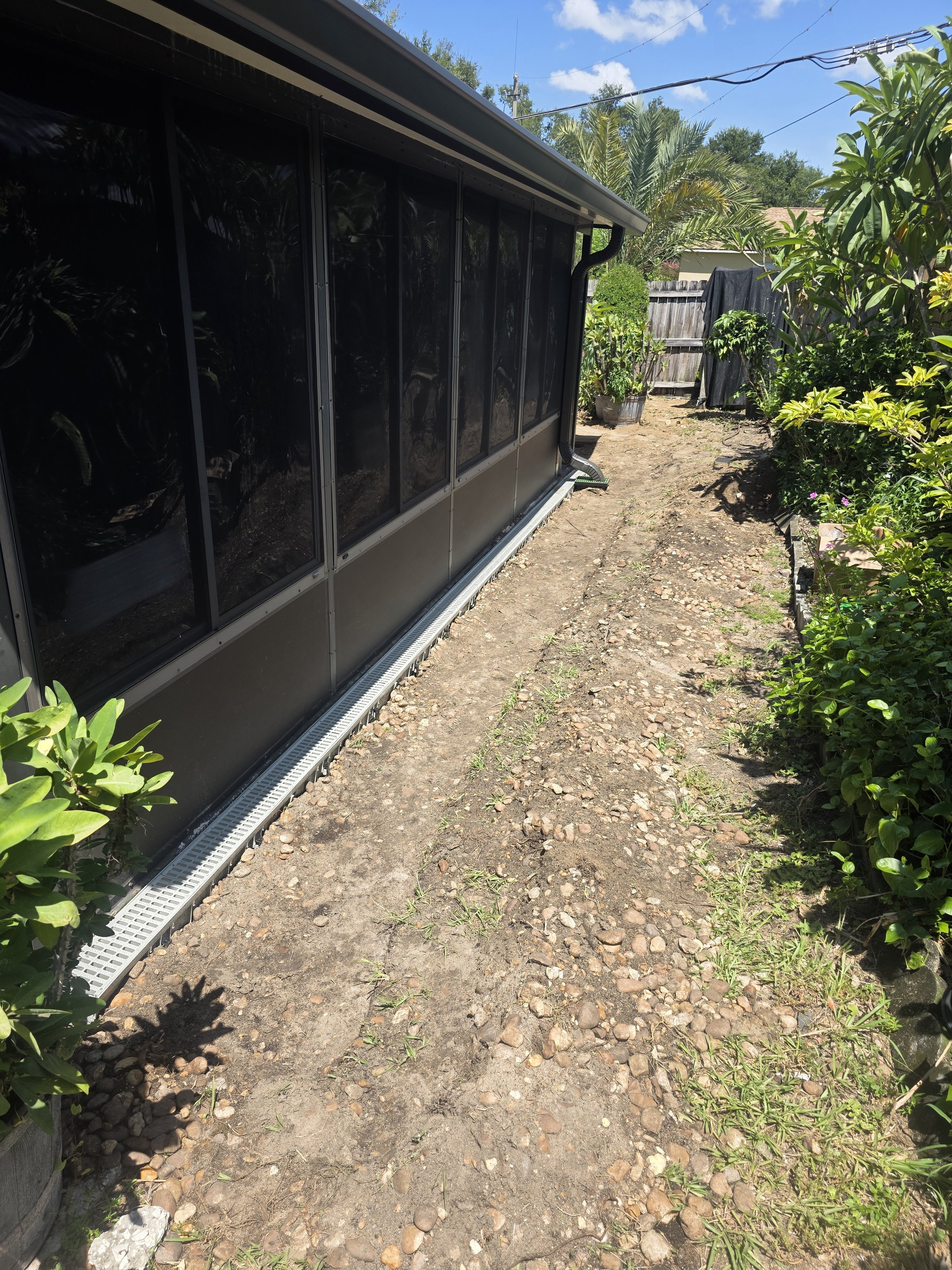 A backyard garden with a gravel path running alongside a black screened porch, surrounded by green bushes and potted plants, with a wooden fence and clear blue sky in the background.