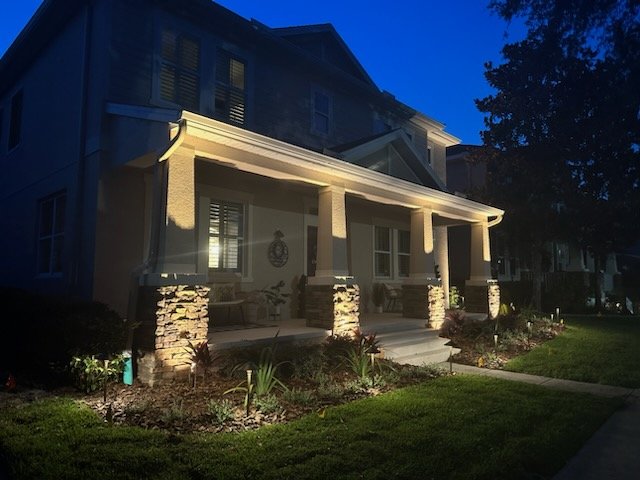 A well-lit house porch at dusk with stone pillars, outdoor seating, and landscaped garden area.